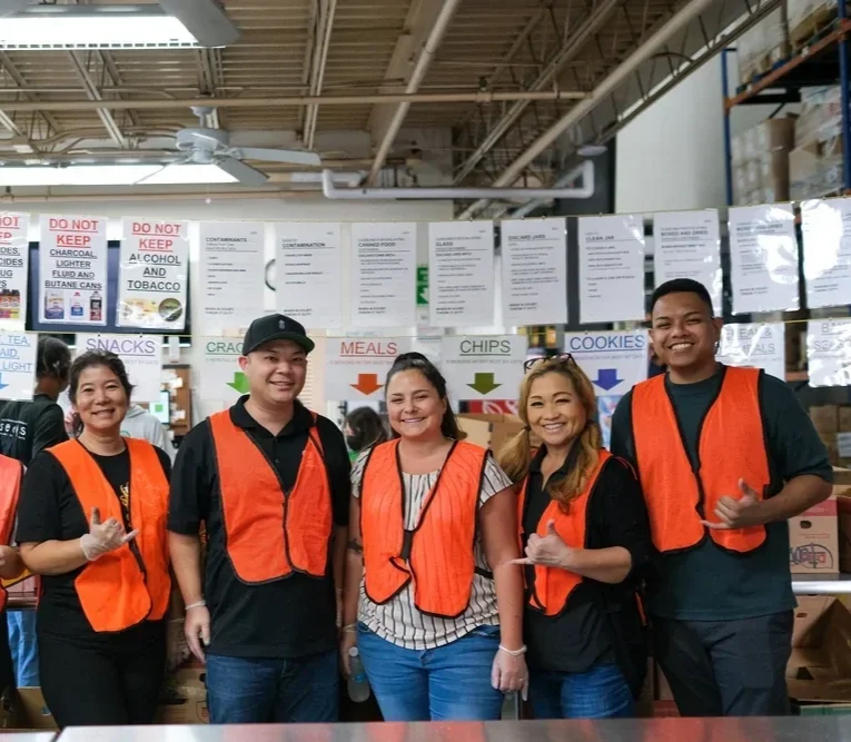 Five volunteers in orange safety vests smiling inside a warehouse, standing in front of shelves labeled 'Snacks,' 'Meals,' 'Chips,' and 'Cookies' with signs about safety regulations in the background.