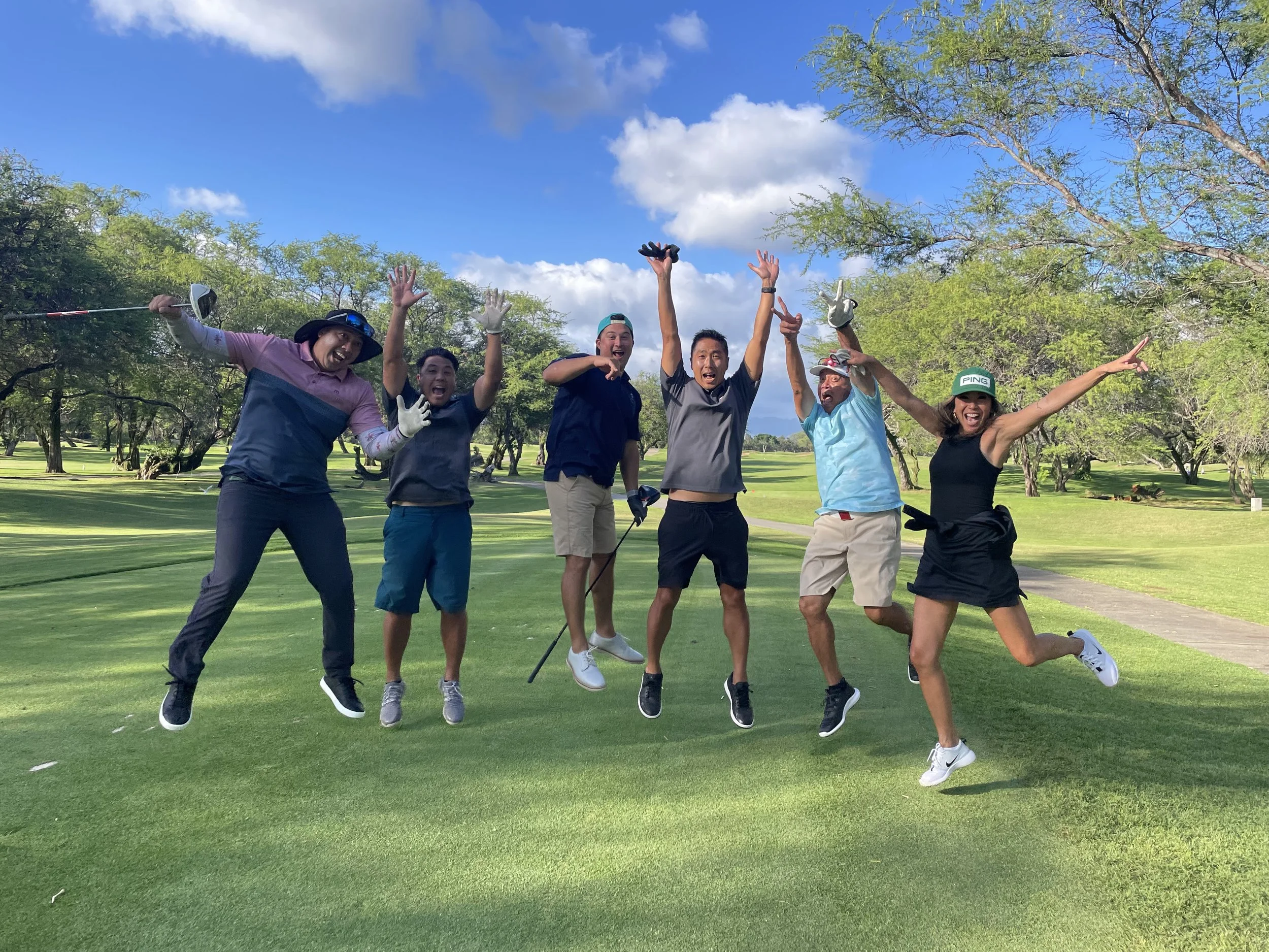 Six people jumping and smiling on a golf course with trees and blue sky in the background.