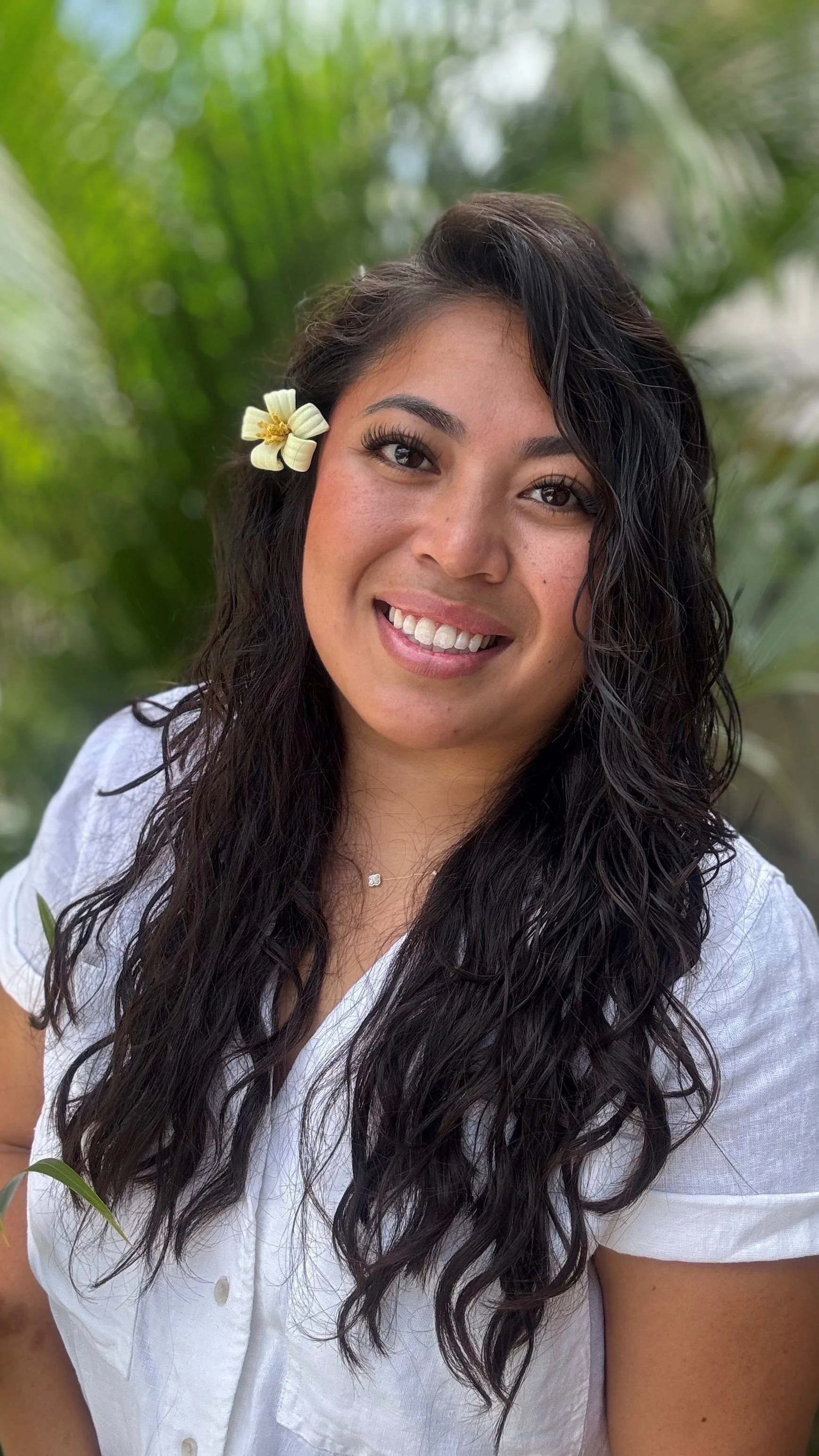 A woman with long curly dark hair, smiling, wearing a white shirt, with a yellow and white flower in her hair, standing outdoors with green plants in the background.
