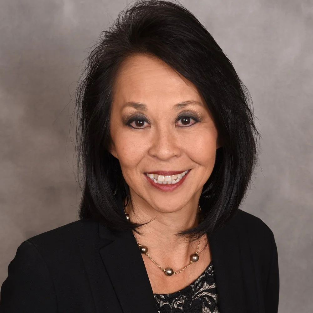 A professional woman with black shoulder-length hair, wearing a black blazer and a pearl necklace, smiling against a gray background.