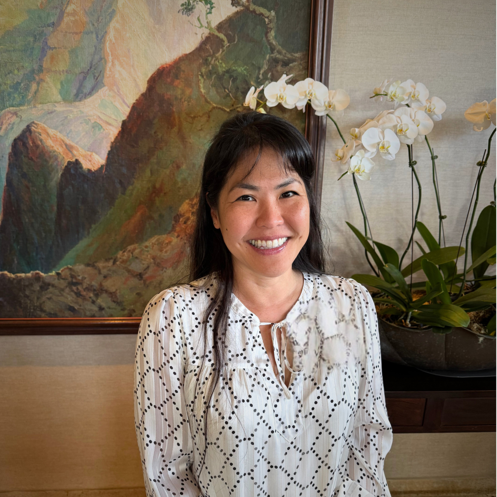 A smiling woman with dark hair standing in front of a framed landscape painting and white orchid plants in a pot.