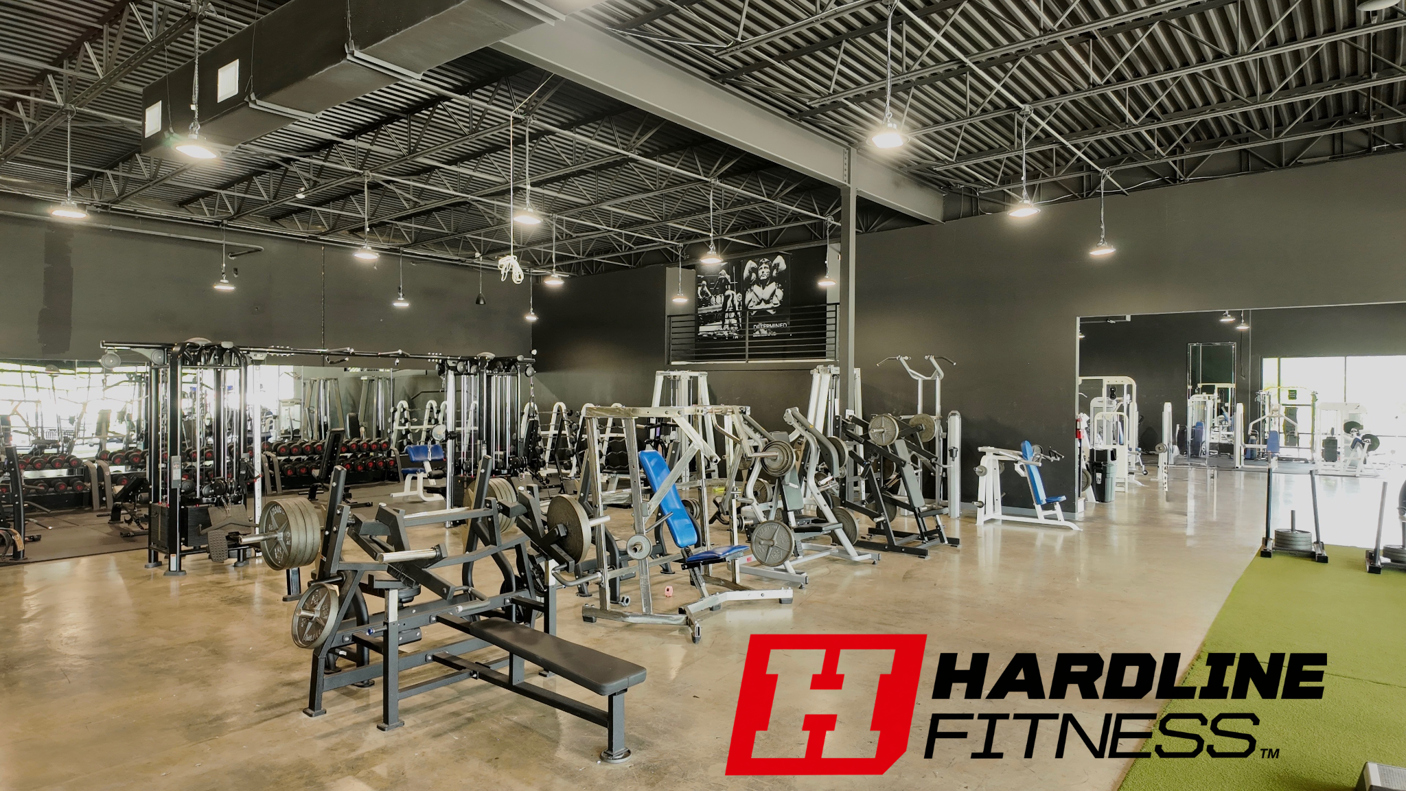 Interior of a gym with various weightlifting equipment and machines, large windows, and a black wall with sports posters.