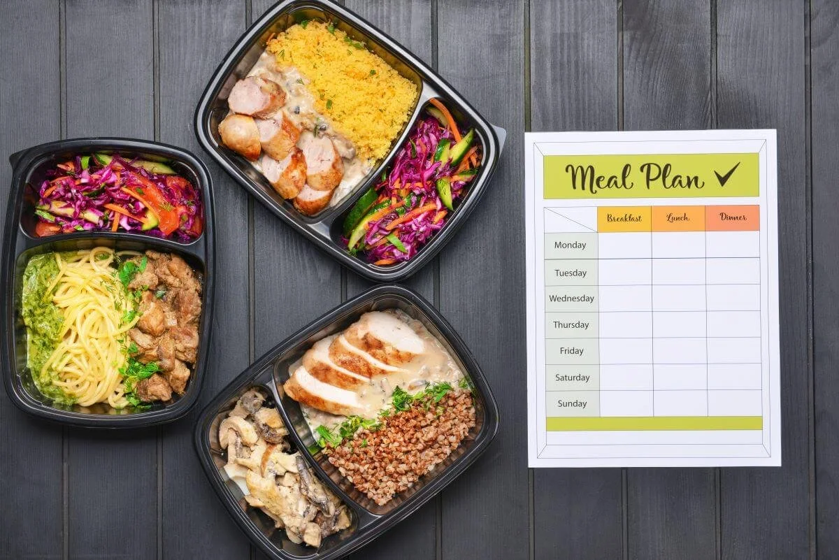 Four meal containers with different foods on a dark wooden table next to a weekly meal plan sheet.