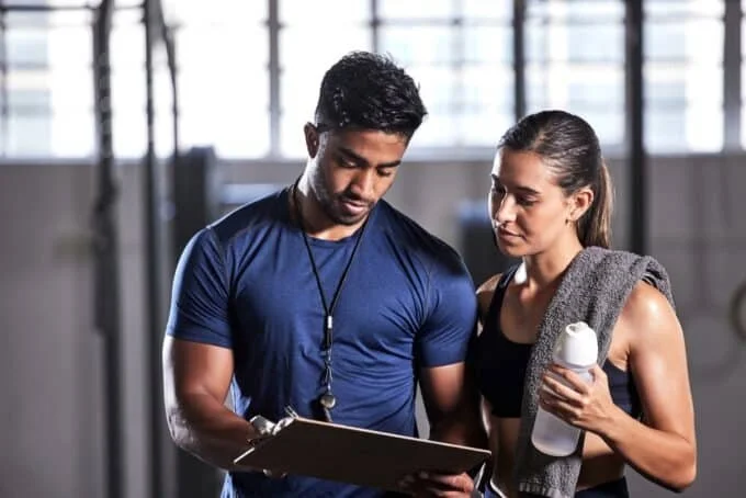 A young man and woman are reviewing a clipboard together in a gym. The man is wearing a blue athletic shirt and a stopwatch around his neck, while the woman is holding a water bottle and has a towel draped over her shoulder.