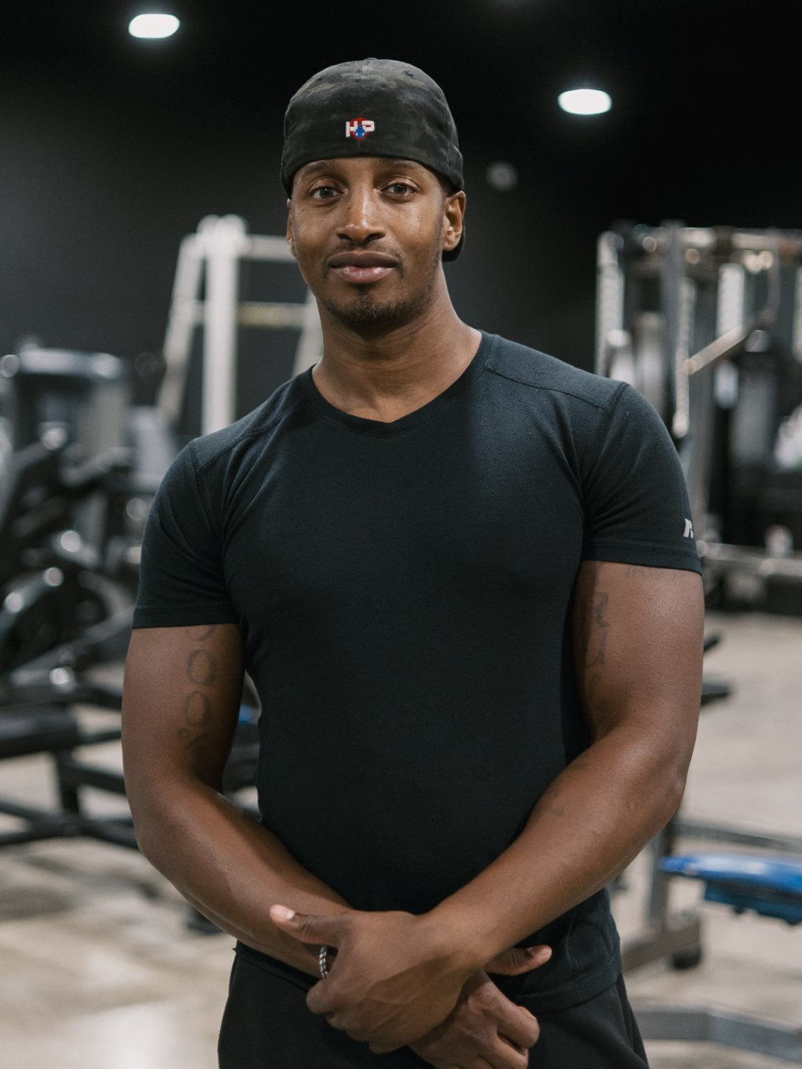 A fit man wearing a black t-shirt and a black cap stands in a gym, with workout equipment visible in the background.