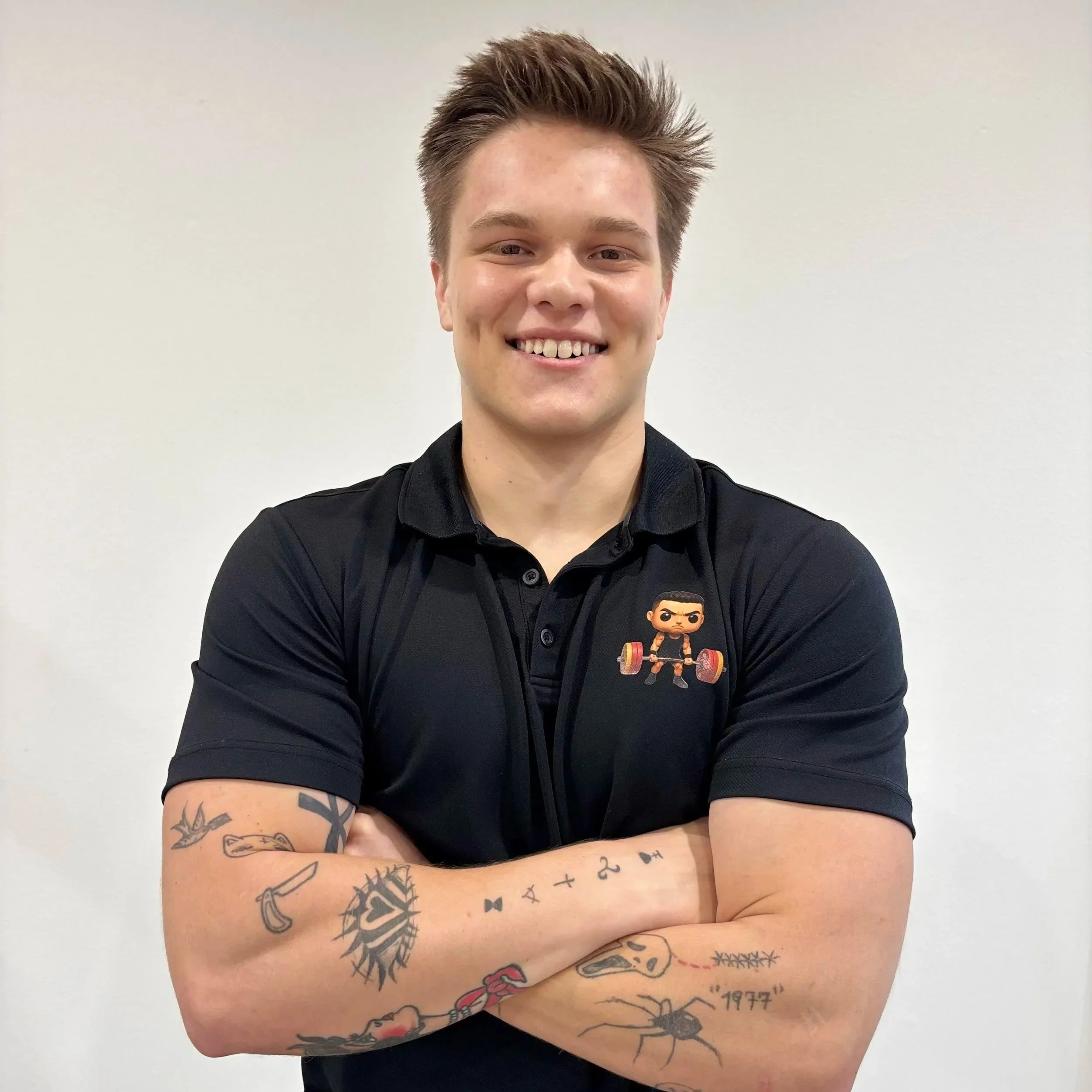 Young man smiling with arms crossed, wearing a black polo shirt with a weightlifting emoji on chest, and displaying various tattoos on both arms against a plain white background.