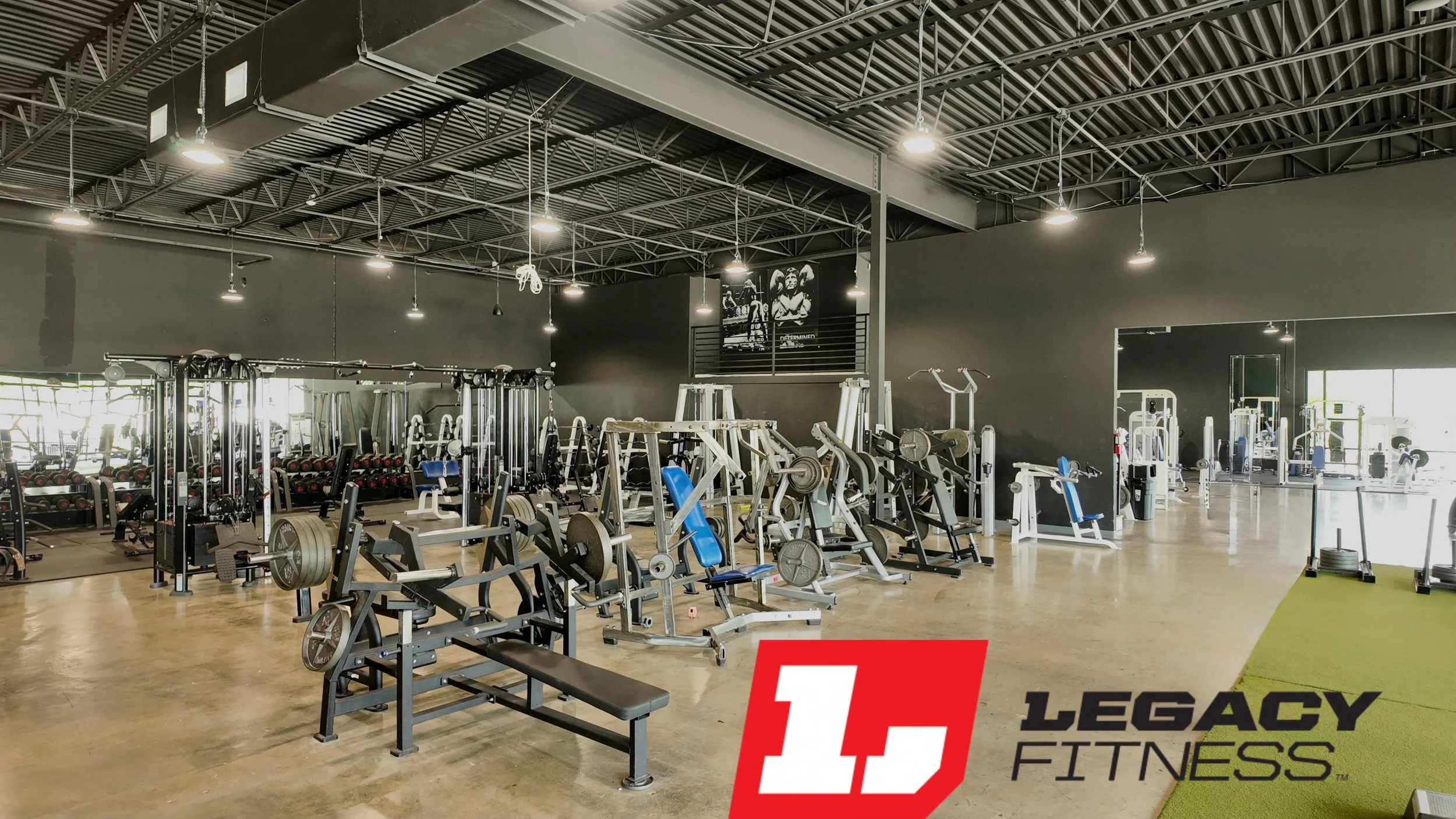 Interior of a modern gym with various weightlifting equipment and machines, hardwood floor, black walls, and a large logo of Legacy Fitness in the bottom right corner.