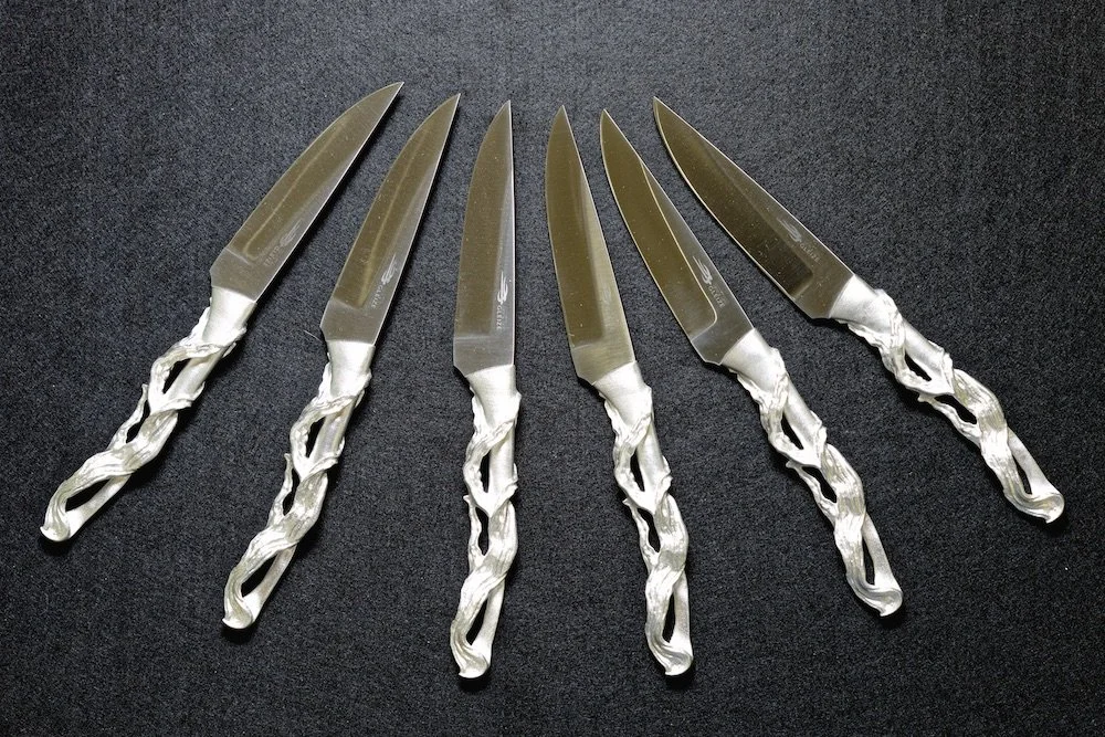 Six kitchen knives with decorative silver handles laid out on a black surface.