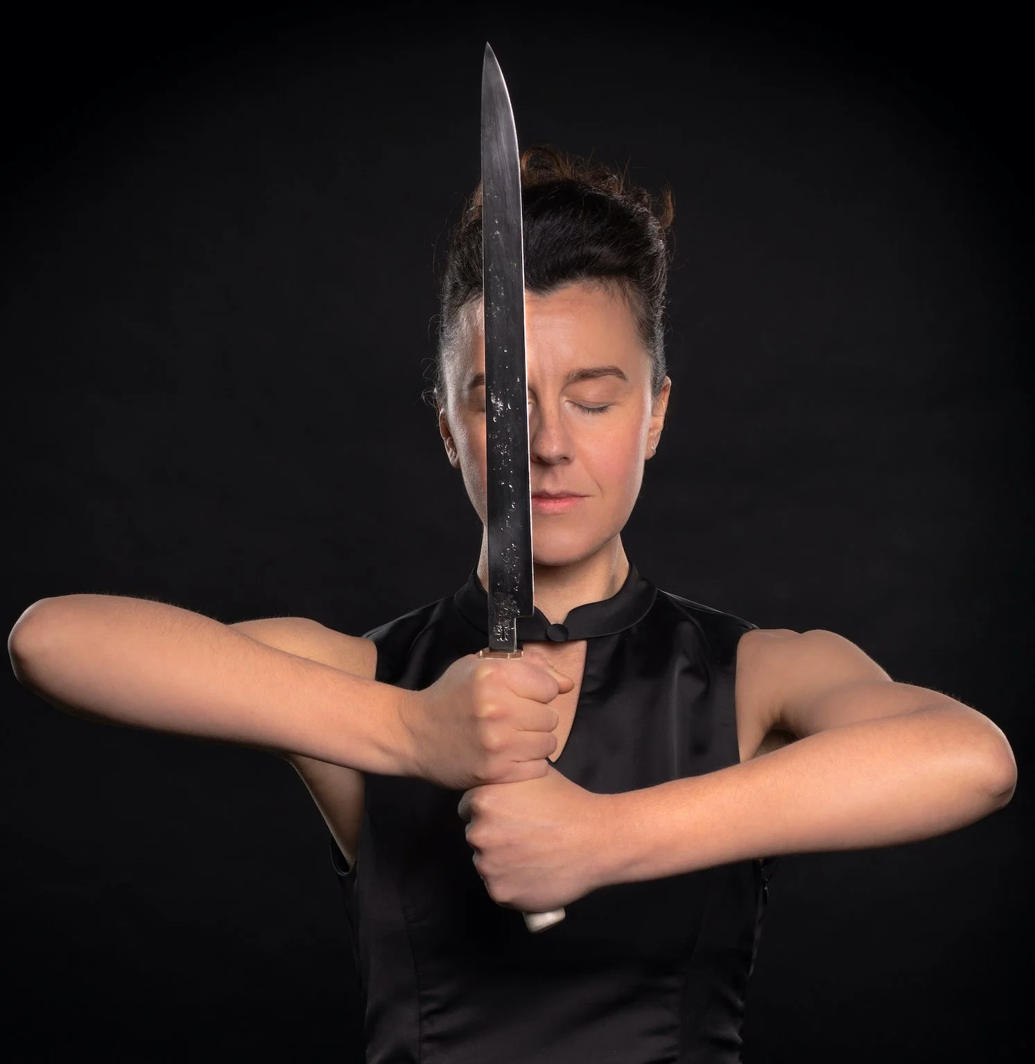 Woman with dark hair tied up, holding a large kitchen knife vertically in front of her face against a black background, eyes closed, wearing a sleeveless black top.