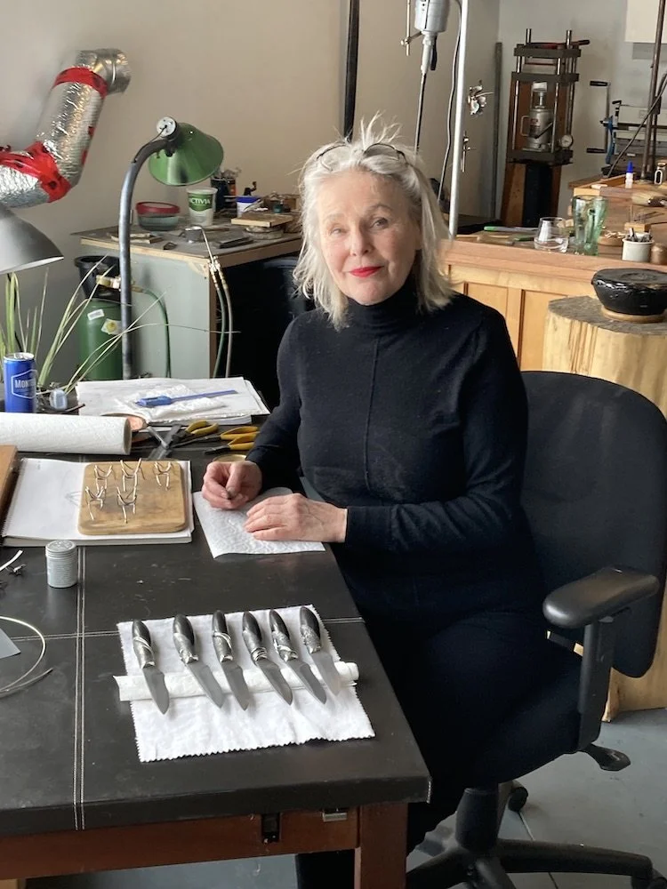A woman with gray hair and red lipstick sitting at a table in a workshop, surrounded by jewelry-making tools, with jewelry pieces laid out on cloths on the table.