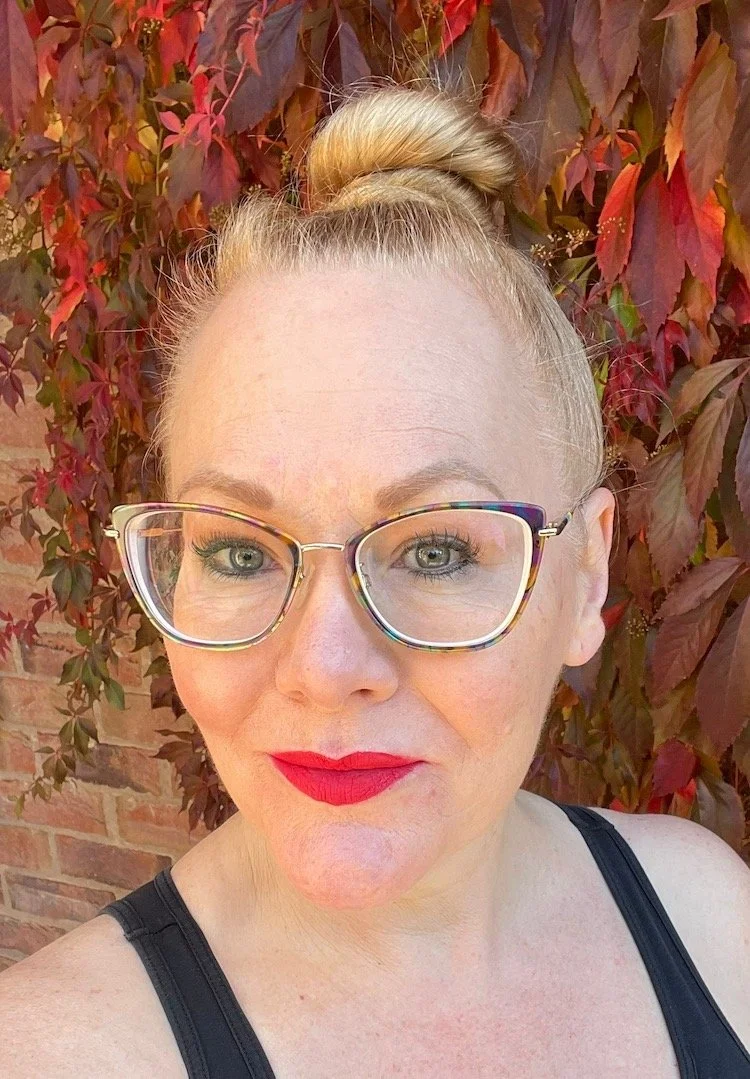 A woman with blonde hair in a bun, wearing glasses with clear frames, red lipstick, and a black tank top, standing outdoors in front of a brick wall covered with red and green leaves.