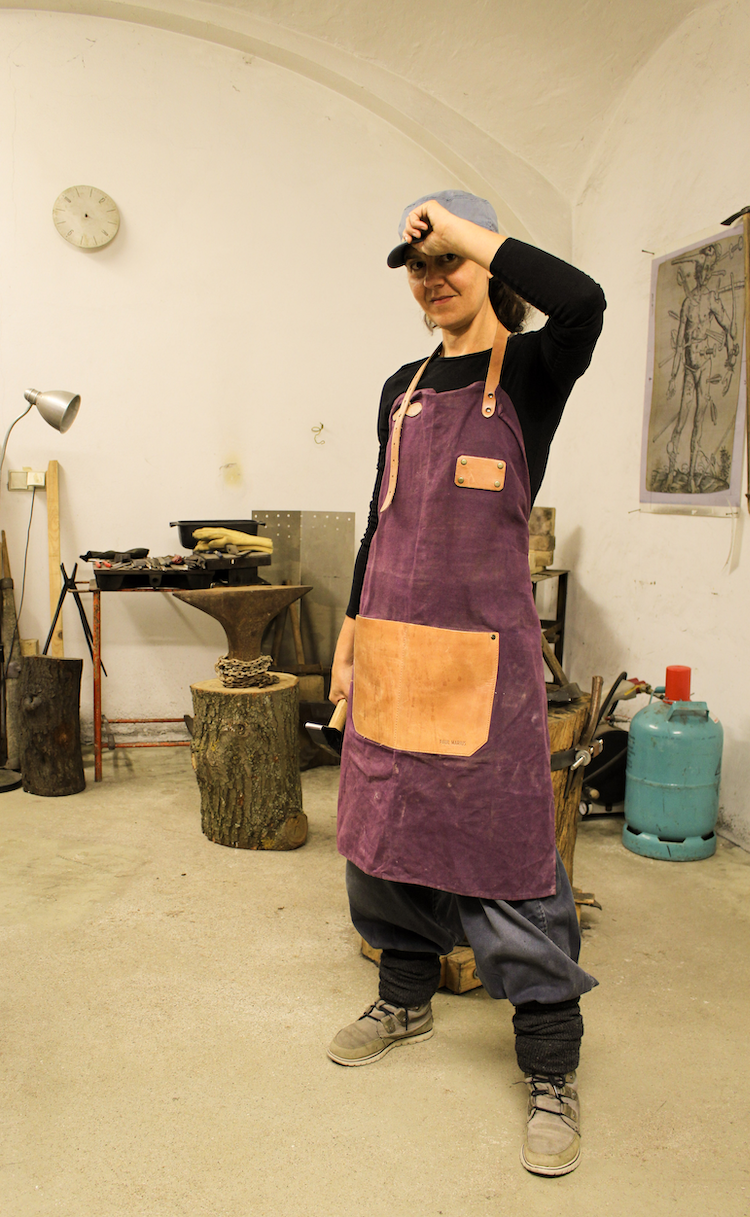 A person in a black shirt, purple apron, and gray cap standing in a woodworking workshop, holding a tool, with tools and wood pieces in the background.