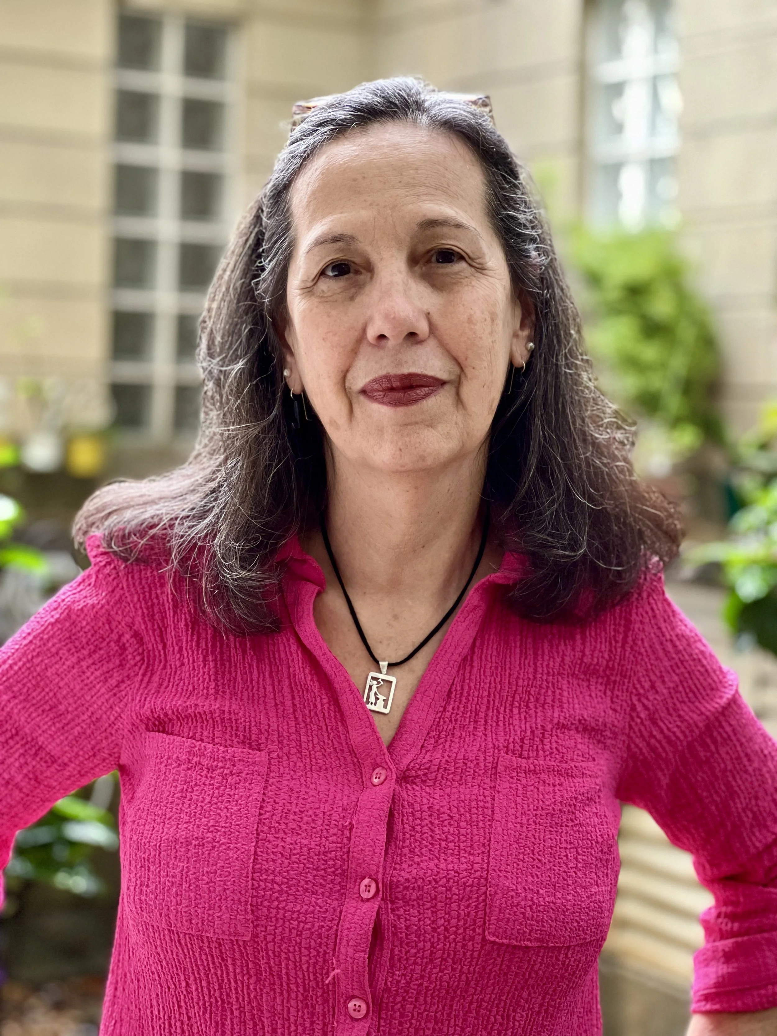 A woman with long dark hair, wearing a pink knit top, earrings, and a necklace, standing outdoors with greenery and a building in the background.