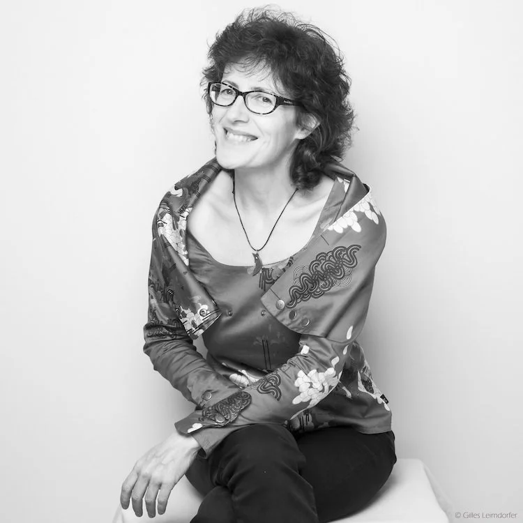 A woman with curly hair and glasses sitting against a plain background, smiling at the camera.