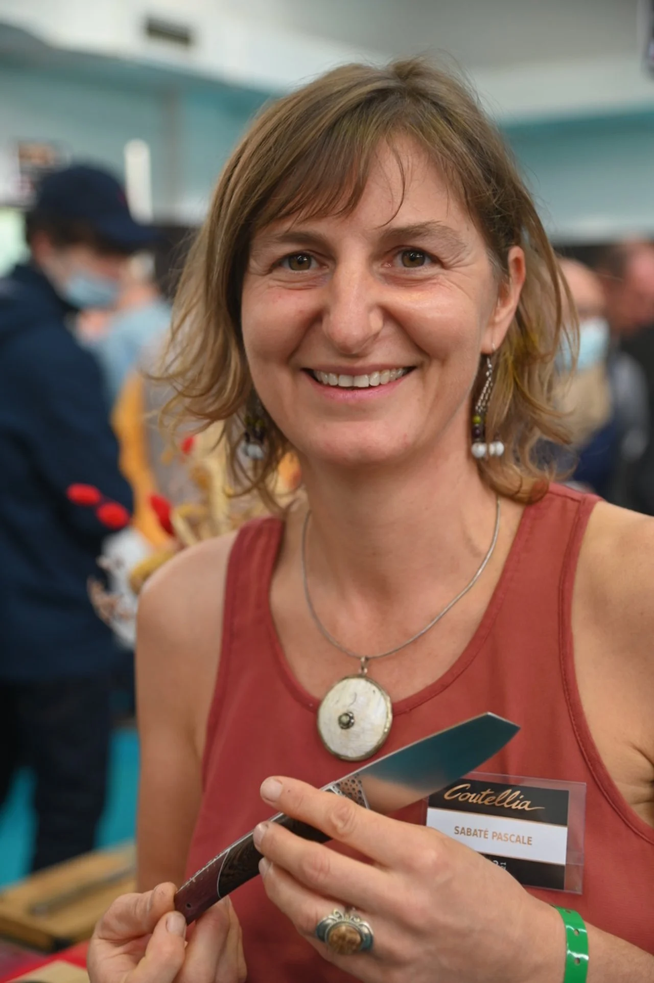 A woman smiling, holding a knife, wearing a red sleeveless top and a name tag that reads 'Sabate Pascale,' with short brown hair and earrings, in a crowded indoor setting.