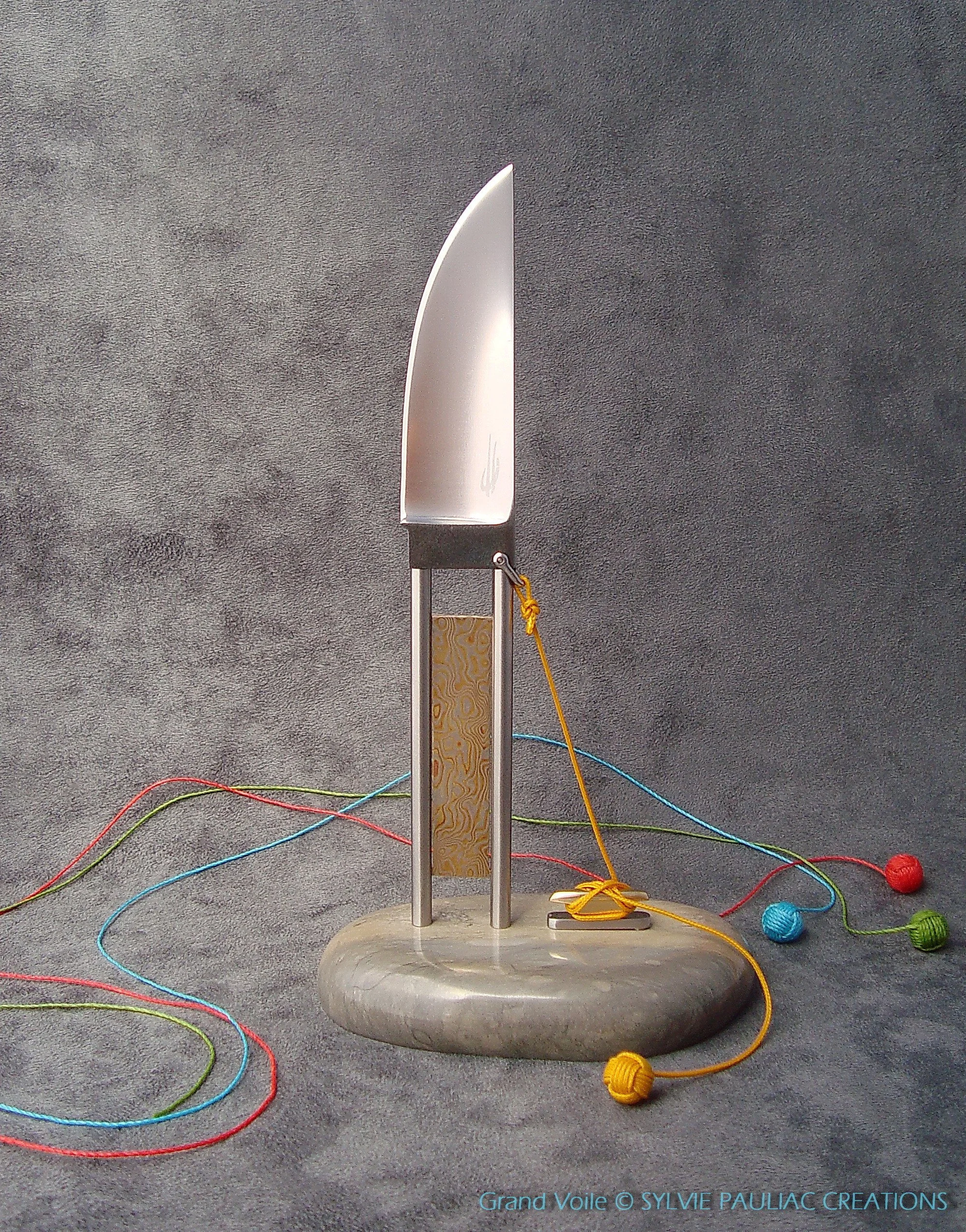 A metallic knife stands upright on a circular gray marble base, surrounded by colorful tangled yarn balls and strings on a gray textured background. The knife is tied to a vertical gray support with yellow string. The artwork is titled 'Grand Voile' by Sylvie Pauliac Creations.