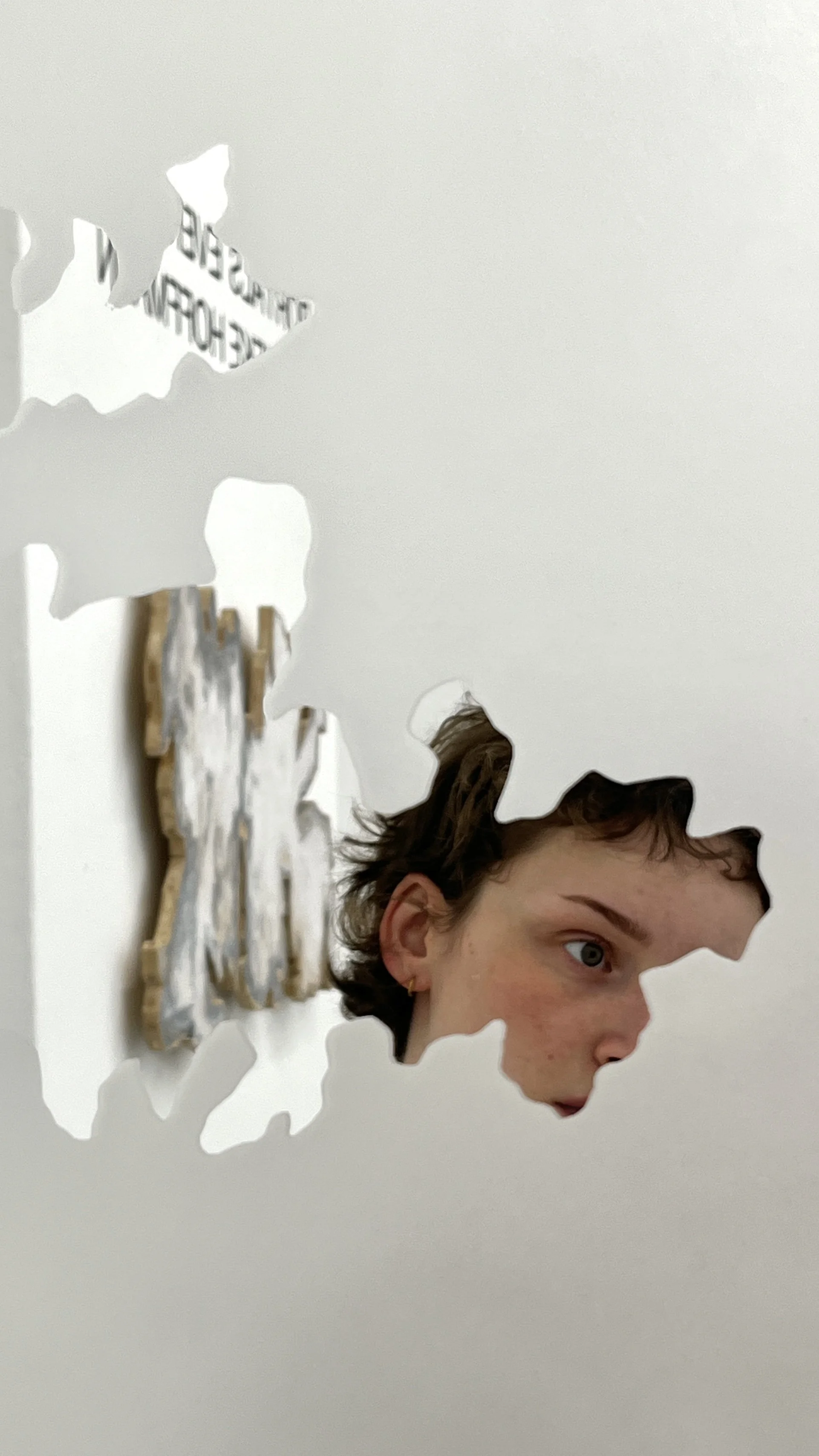 Person's face seen through a cut-out in a white wall, creating a mirror effect.
