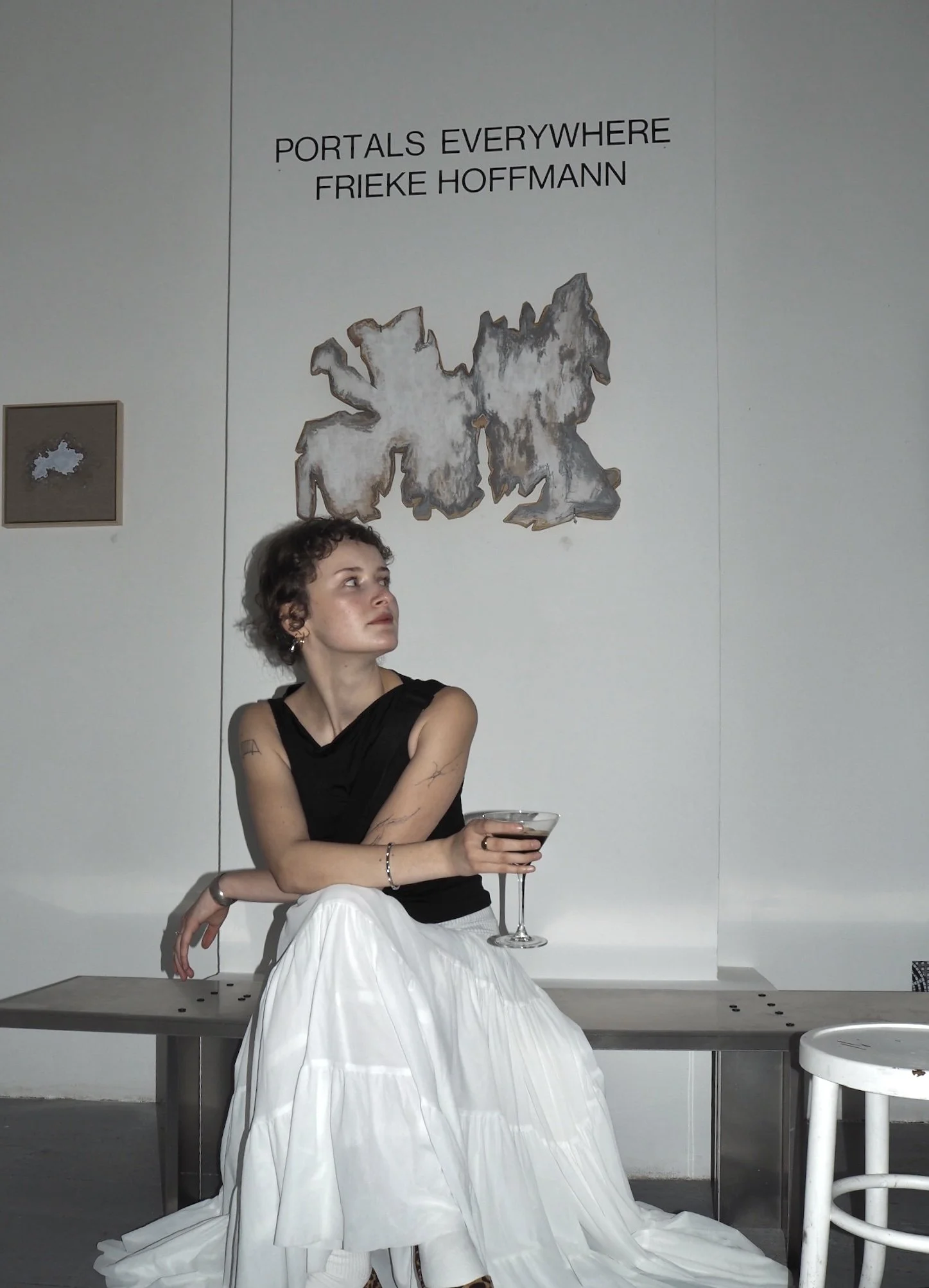 A woman with short curly hair and tattoos on her arms sitting on a bench, holding a cocktail glass, with art and text on the wall behind her at an art exhibition.