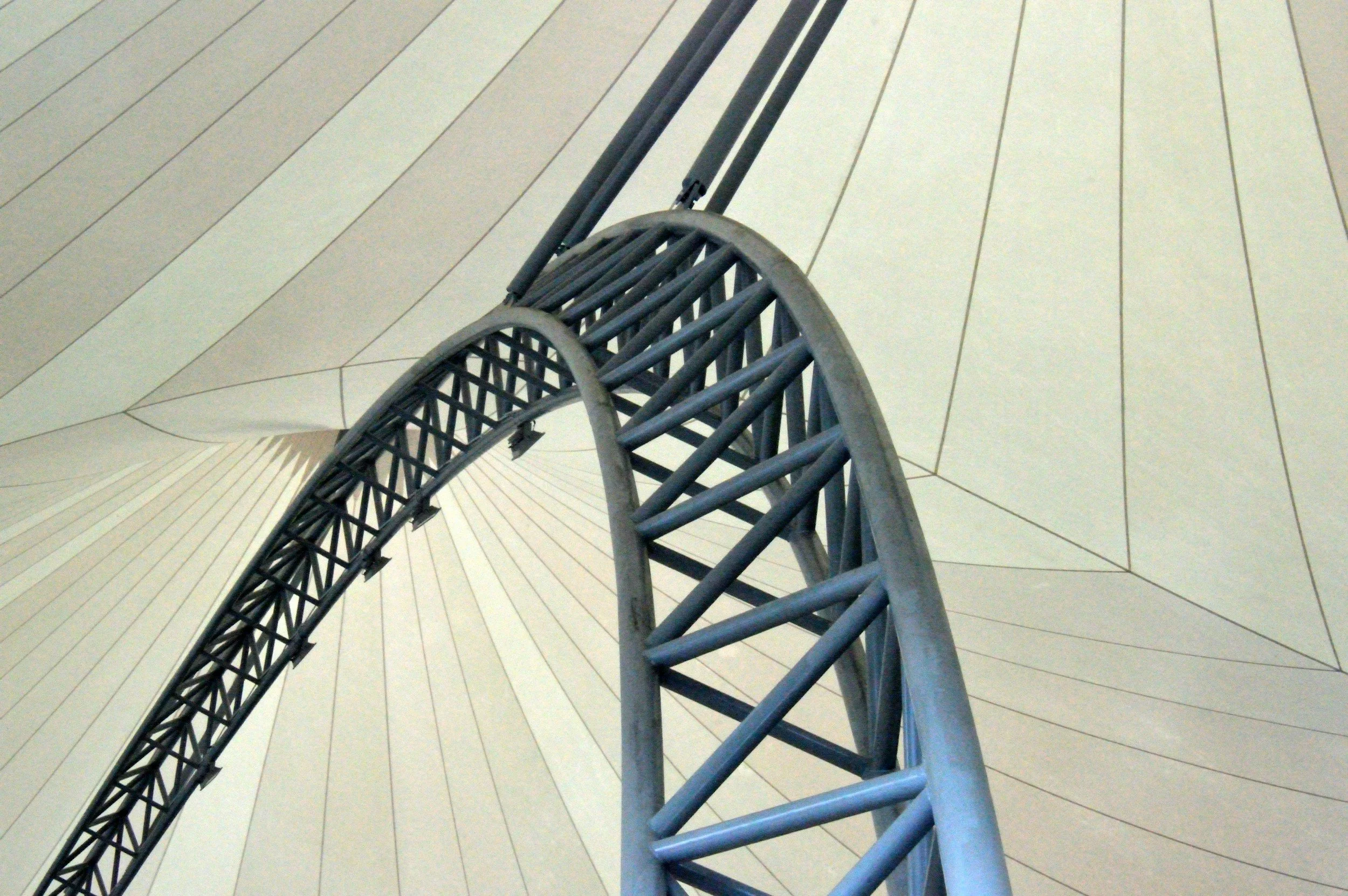 Close-up view of a roller coaster track inside a large sheltered space with a metallic structural framework supporting a fabric roof, taken from below.