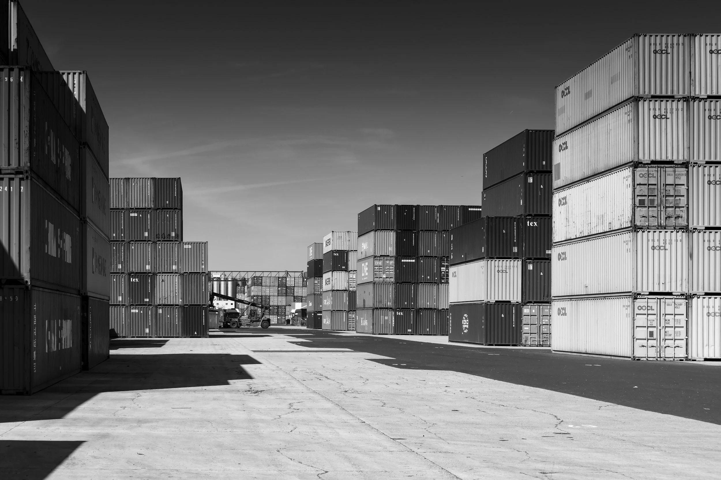 A shipping yard with numerous stacked shipping containers, a forklift, and a tractor under a clear sky.