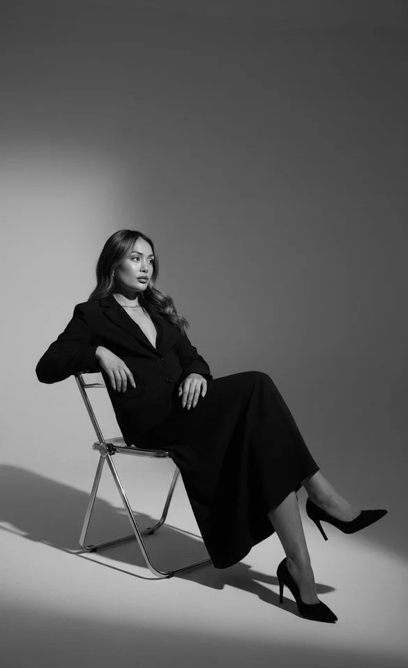 Black and white photo of a woman in a dark suit and high heels sitting on a chair with a minimalist background.