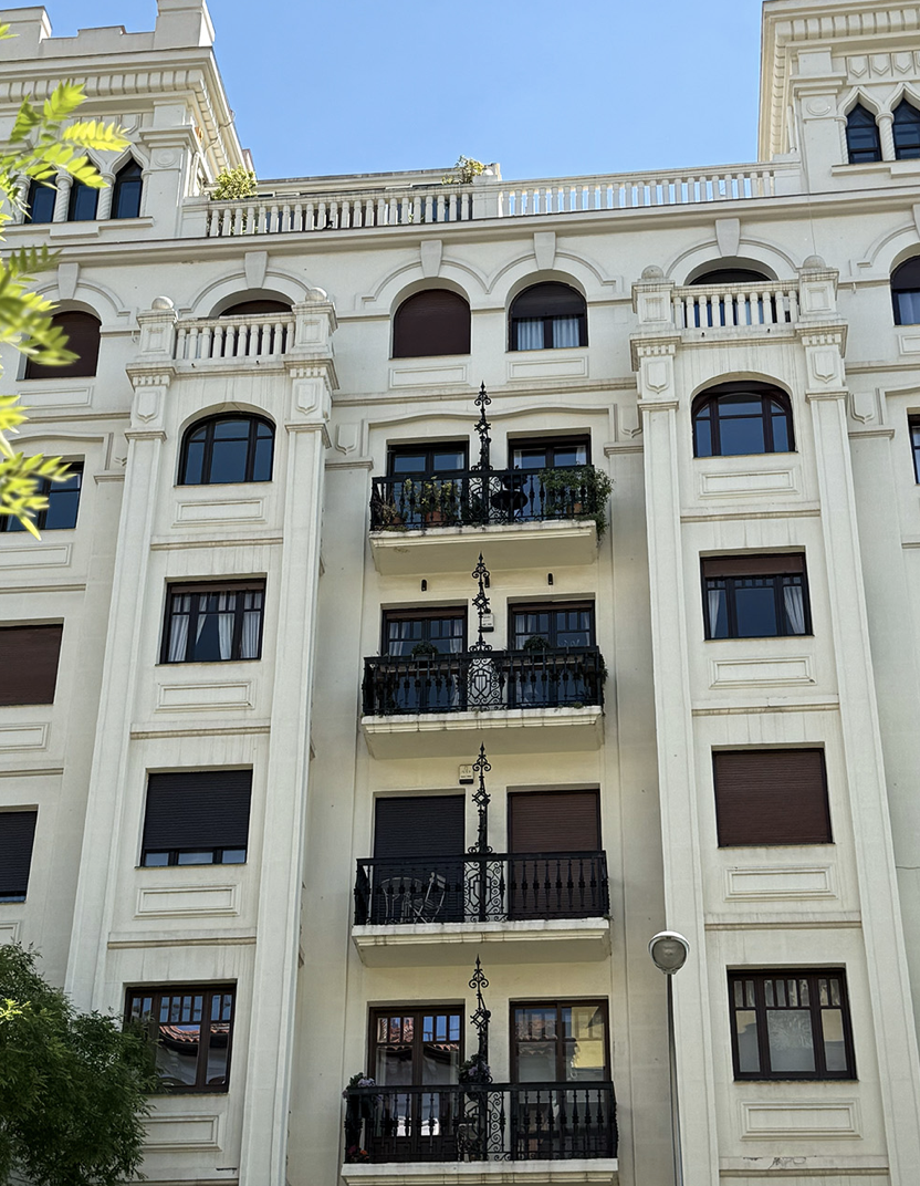 Edificio de varios pisos con balcones de hierro forjado, ventanas con contraventanas y detalles arquitectónicos en color blanco.