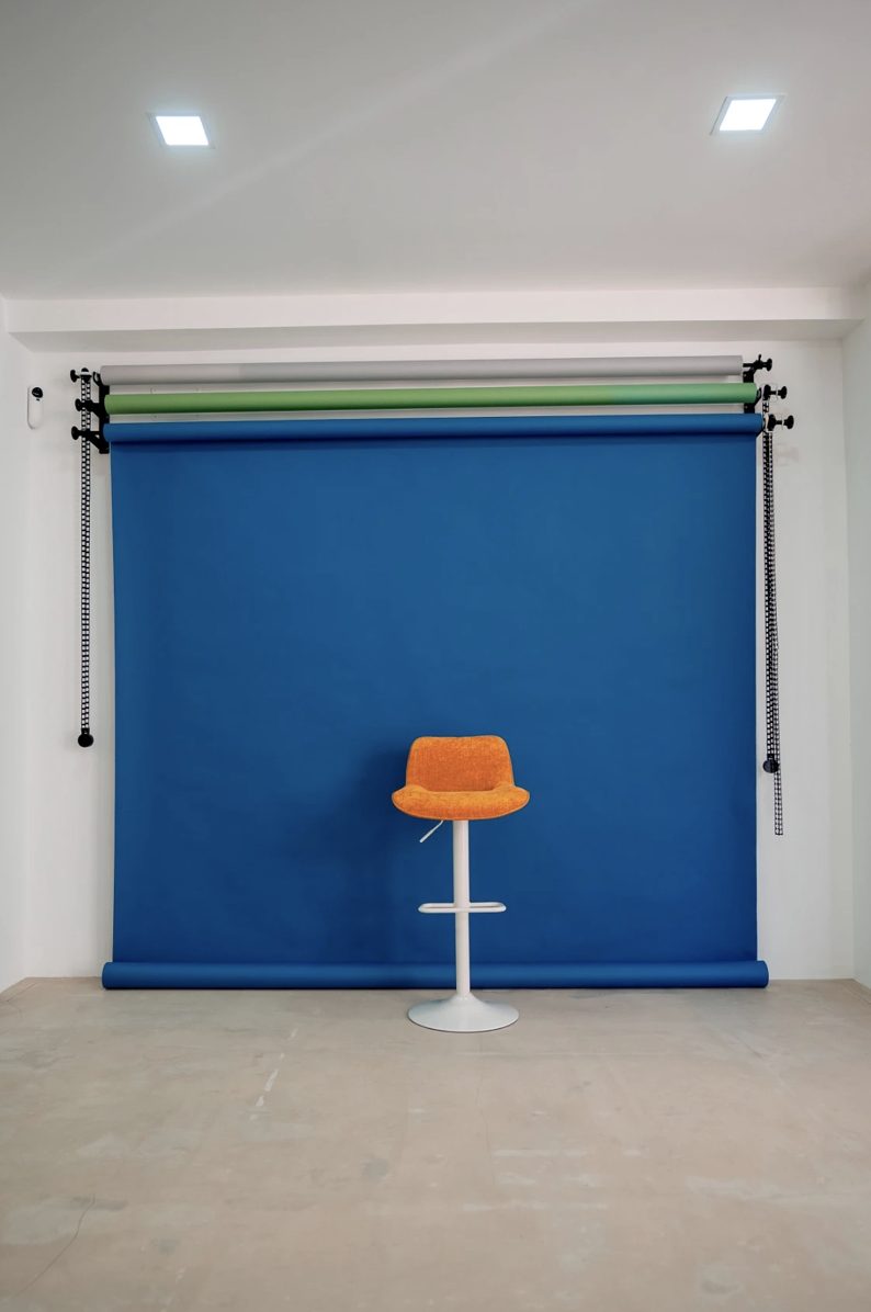 Fondo azul con una silla naranja y un fondo de tela para fotografía, en un espacio con paredes blancas y techo con luces empotradas.