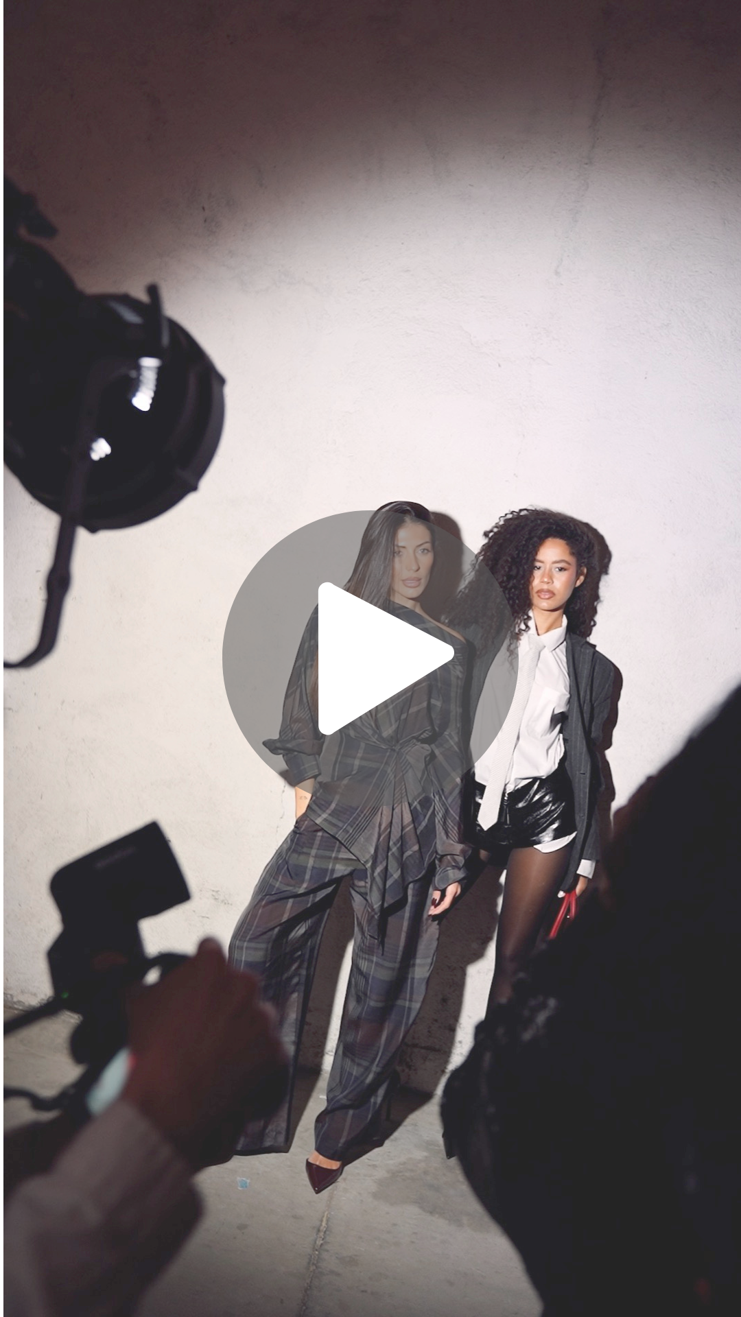 Dos modelos posando para la sesión de fotos, una con pantalones a cuadros y otra con short de cuero y camisa blanca, en un estudio con fondo blanco.