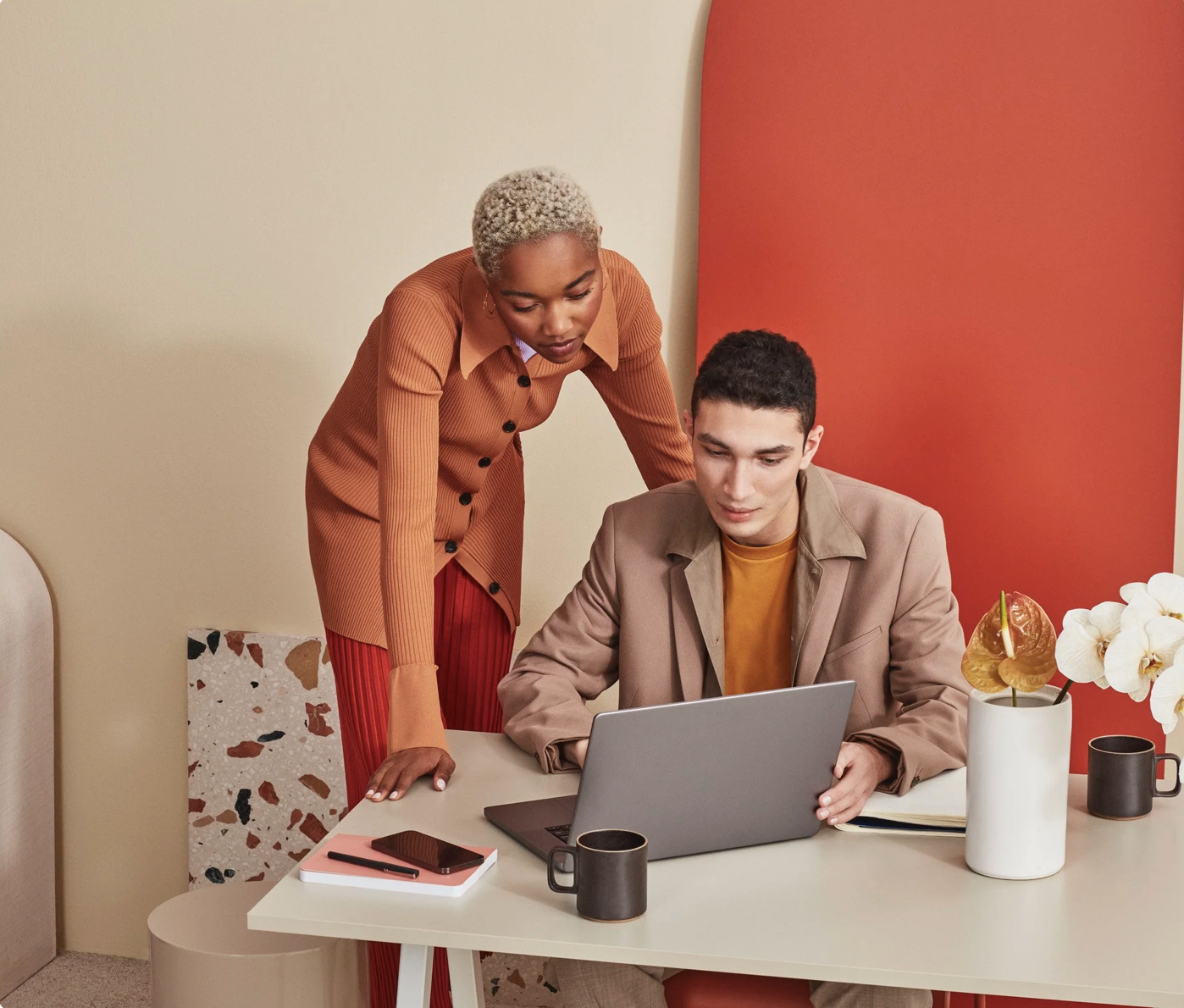 Una mujer y un hombre trabajan en una oficina, la mujer está de pie y aparece mirando la pantalla de una computadora portátil que el hombre tiene frente a él, en una mesa blanca con dos tazas negras, un libro, una libreta y una planta con flores blancas.