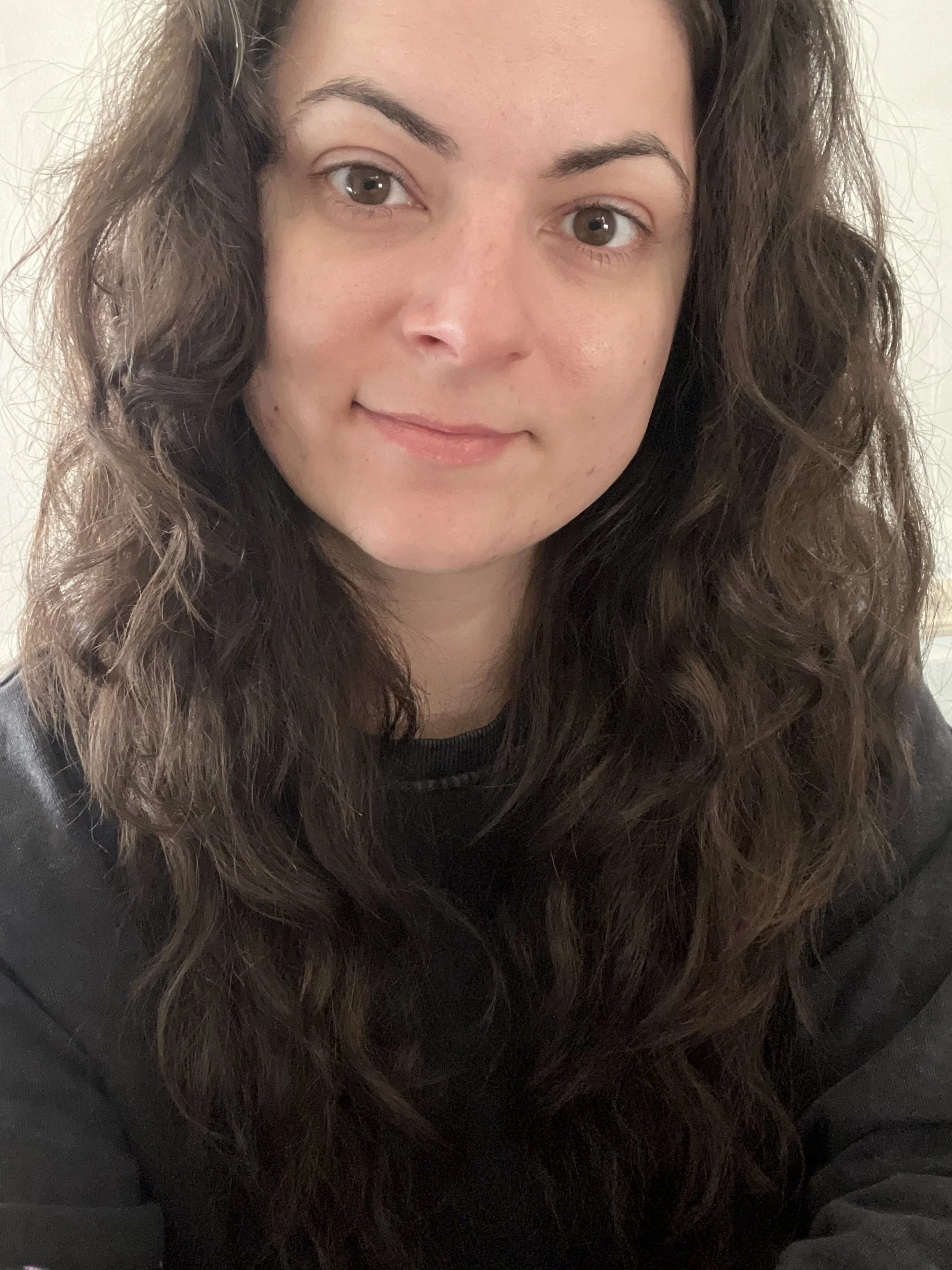 Close-up photo of a woman with long, dark brown curly hair, brown eyes, and light skin, wearing a black top and a faint smile.