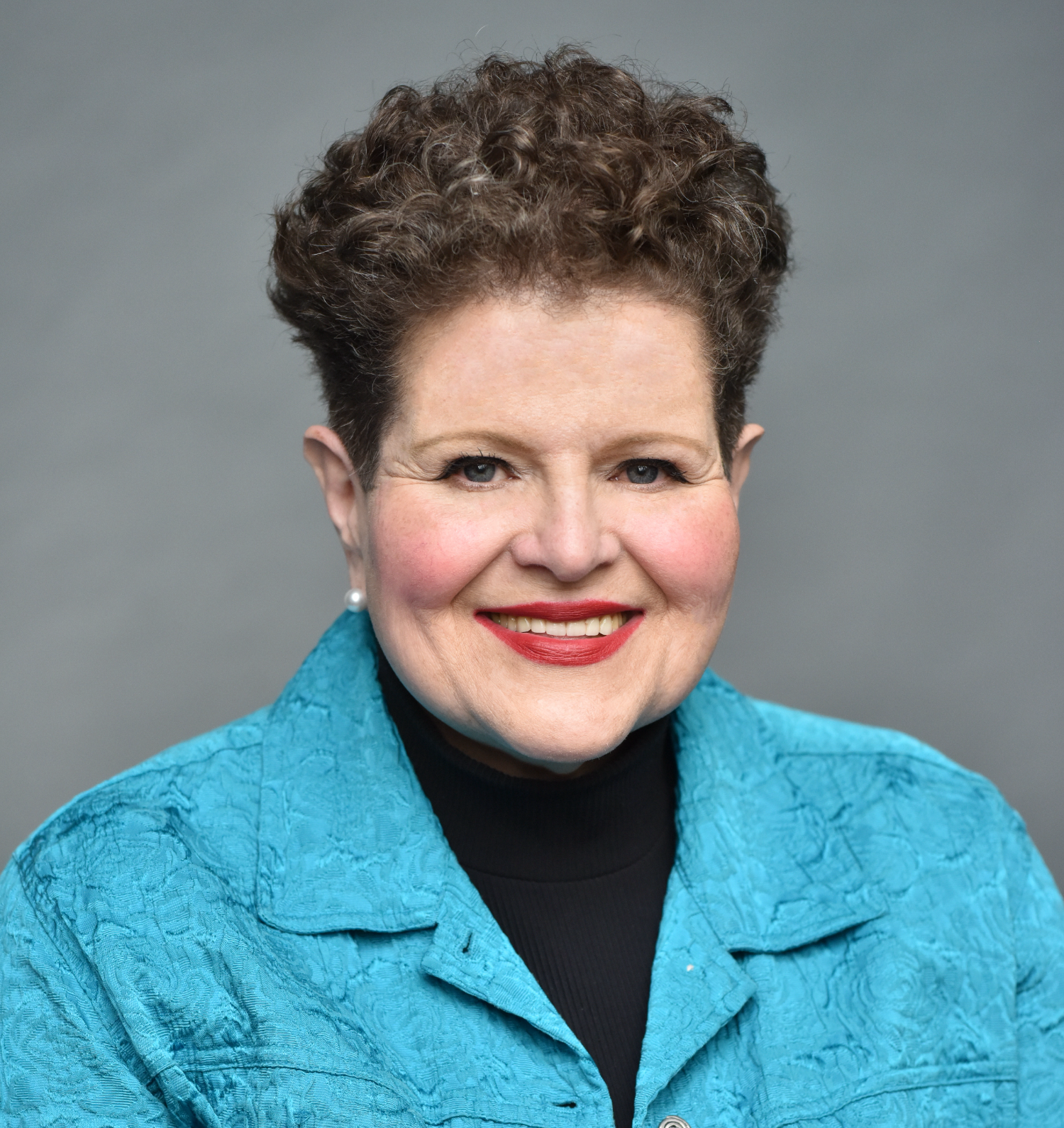 A professional headshot of a woman with short, curly brown hair, wearing a turquoise textured blazer over a black turtleneck, and pearl earrings, smiling against a gray background.