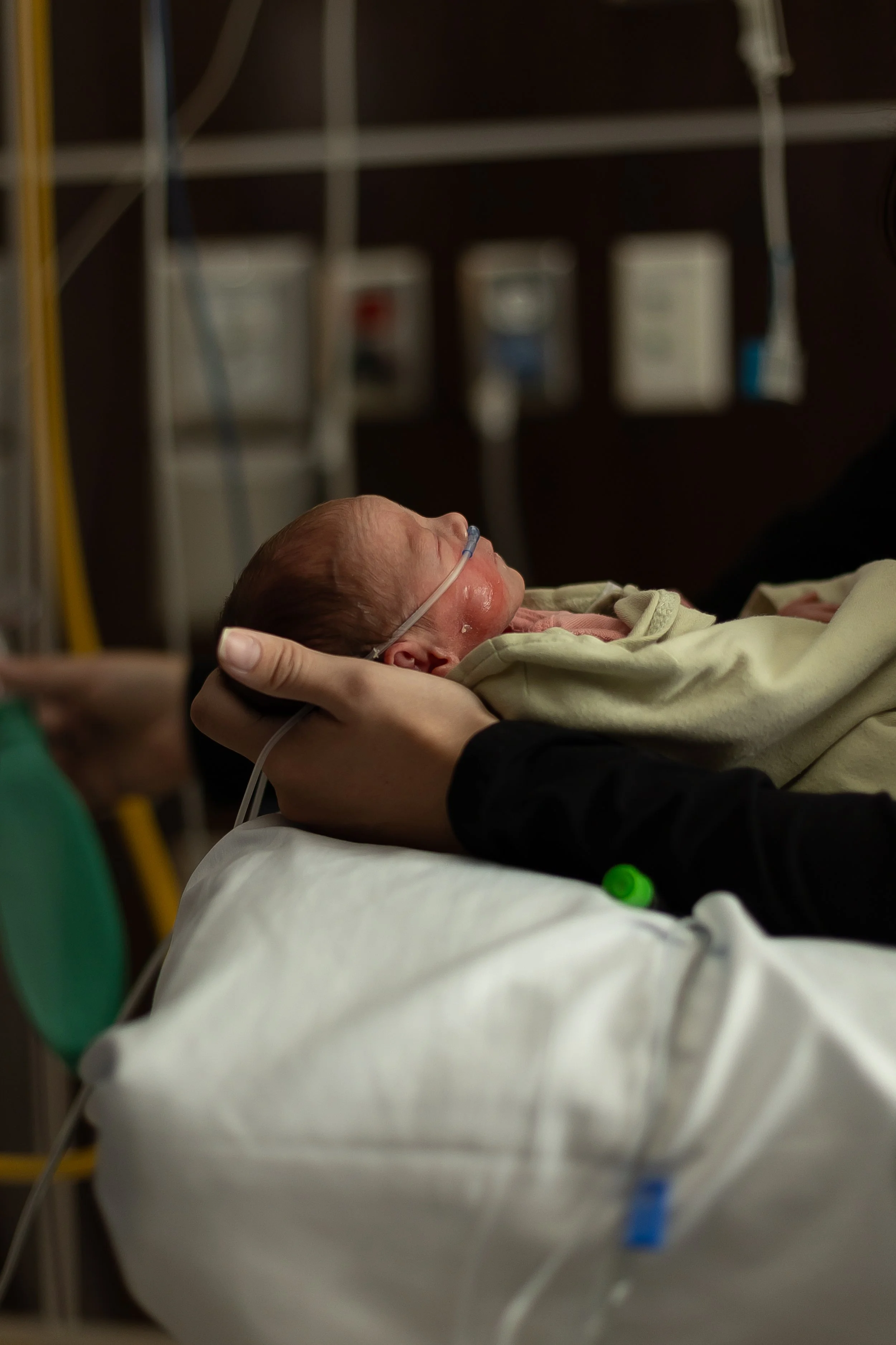 A newborn baby with a nasal cannula, lying on a hospital bed being gently held by an adult.