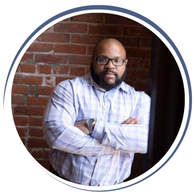 A man with glasses and a beard, wearing a light plaid shirt, standing with arms crossed in front of a brick wall.