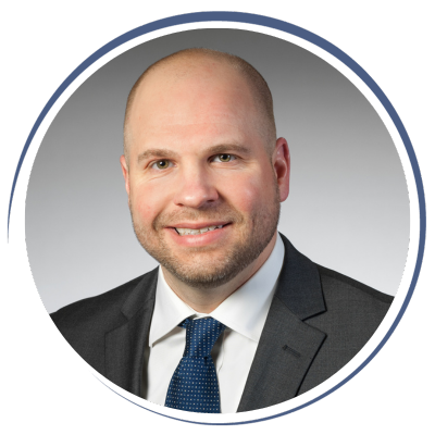 A headshot of a bald man wearing a suit and tie, smiling against a plain gray background.