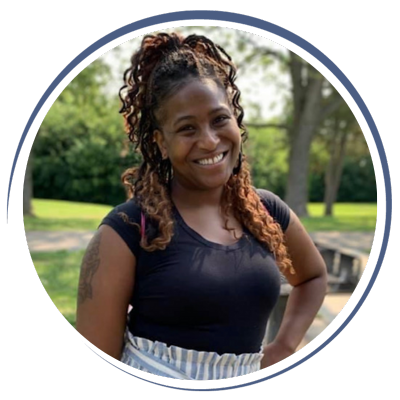 A woman with curly hair smiling outdoors in a park, wearing a black shirt and striped skirt.