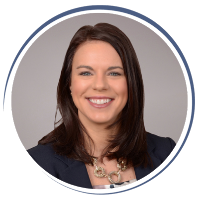 A smiling woman with shoulder-length brown hair wearing a dark blazer and a chunky necklace against a neutral background.