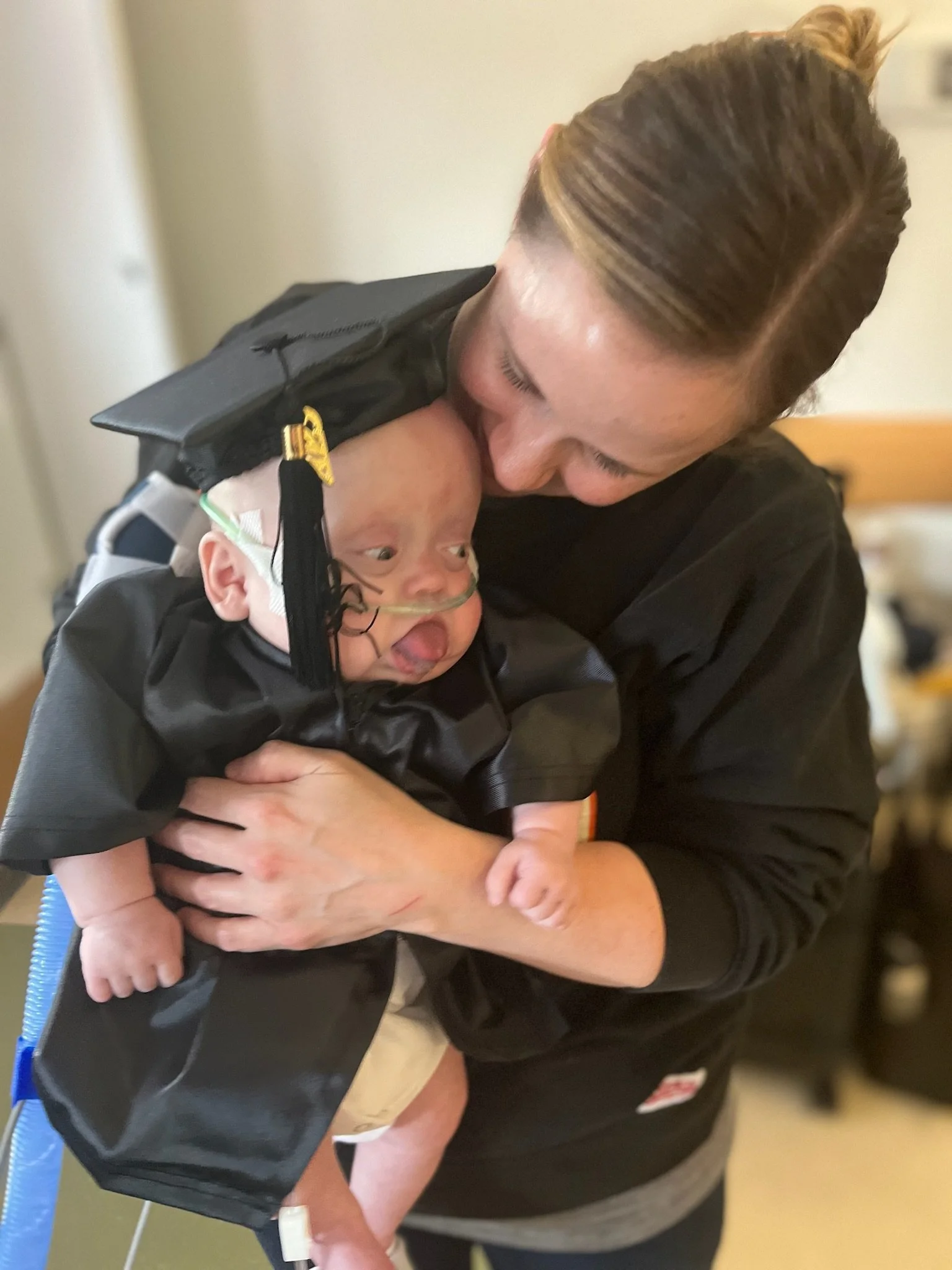 A woman in a black jacket kisses a baby with medical tube and oxygen mask, who is being held close.