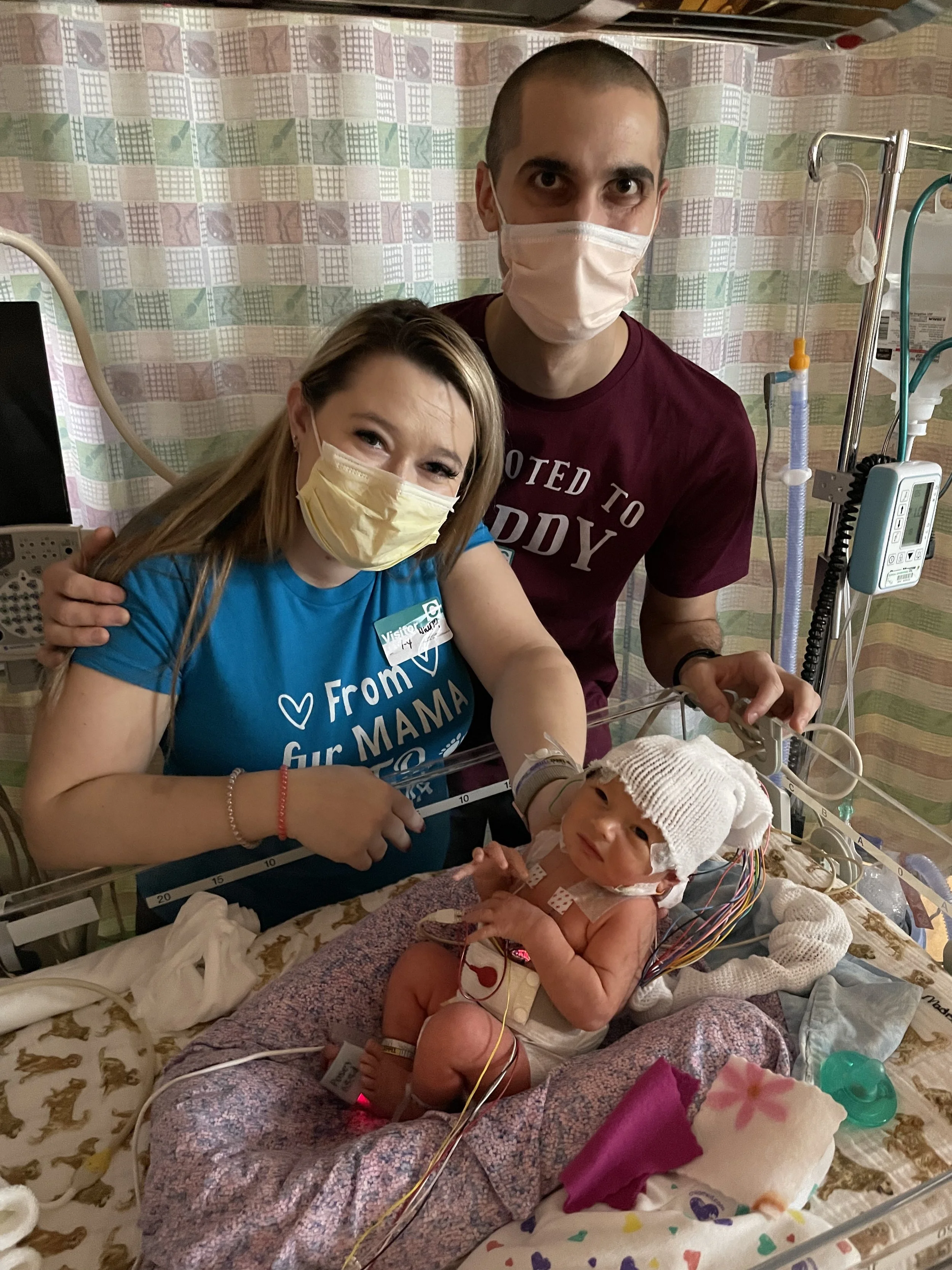 A woman, man, and a baby in a hospital room, all wearing masks. The woman and man are smiling and looking at the camera, with the woman sitting next to the baby, who is in an incubator with medical wires and equipment.