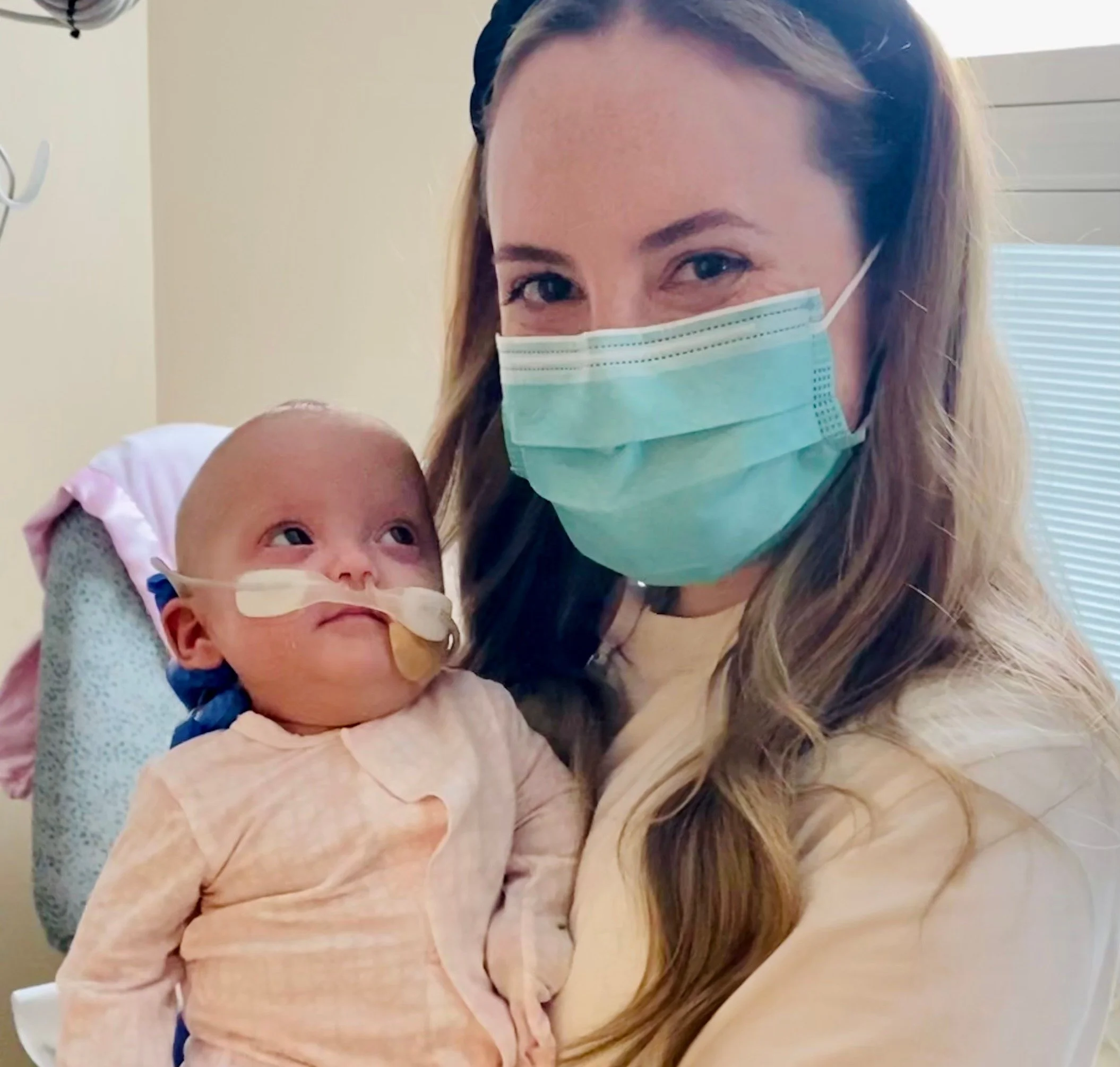 A woman wearing a surgical mask holding a young child in a hospital setting.