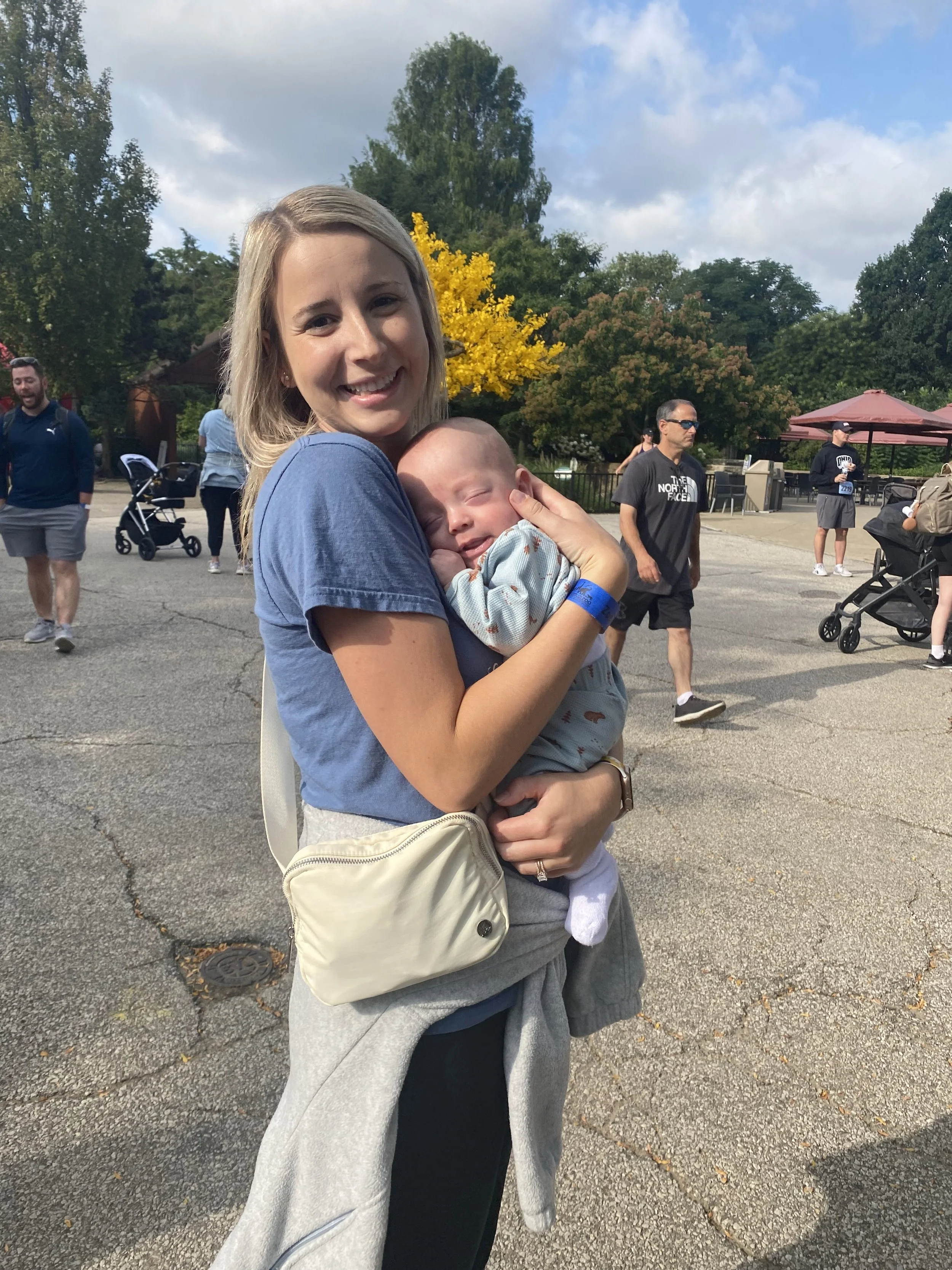 A smiling woman holding a sleeping baby in a crowded outdoor setting with trees and people in the background.