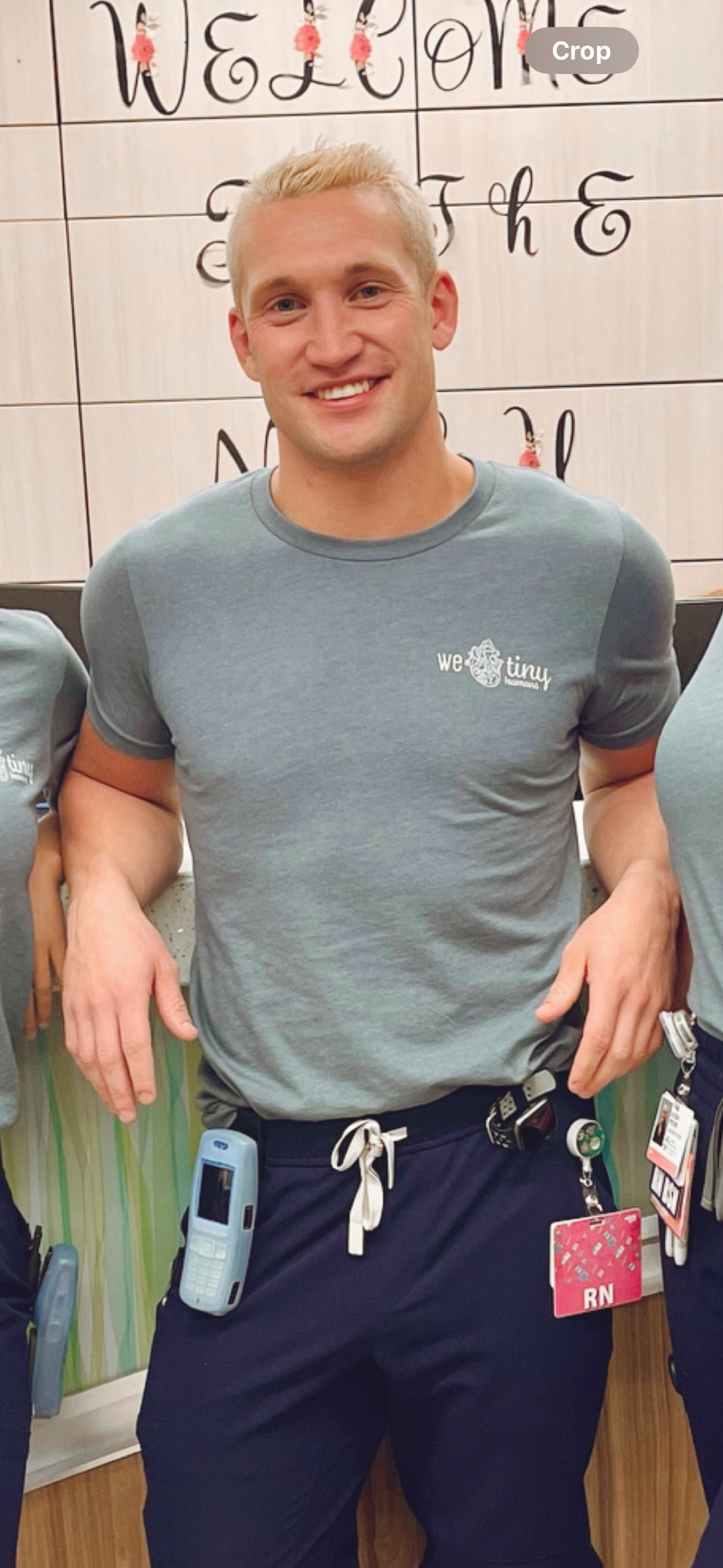 A young man with blond hair smiling in a healthcare setting, wearing a gray T-shirt with a logo that reads 'we tiny' and dark pants with a clip-on ID badge, a walkie-talkie, and a cellphone attached to his waistband.