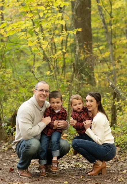 A family of four outdoors in a wooded area during fall, with yellow and green leaves, posing for a photo. The father and mother are kneeling, each holding a young child, all smiling at the camera.