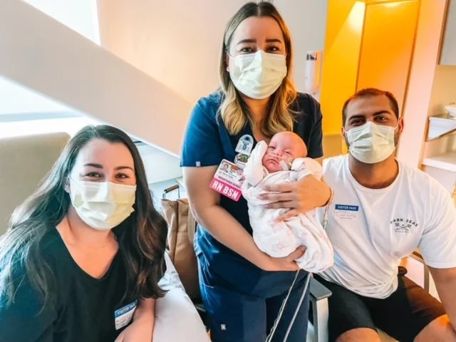 A healthcare team with two women, a man, and a newborn baby all wearing face masks in a hospital room.