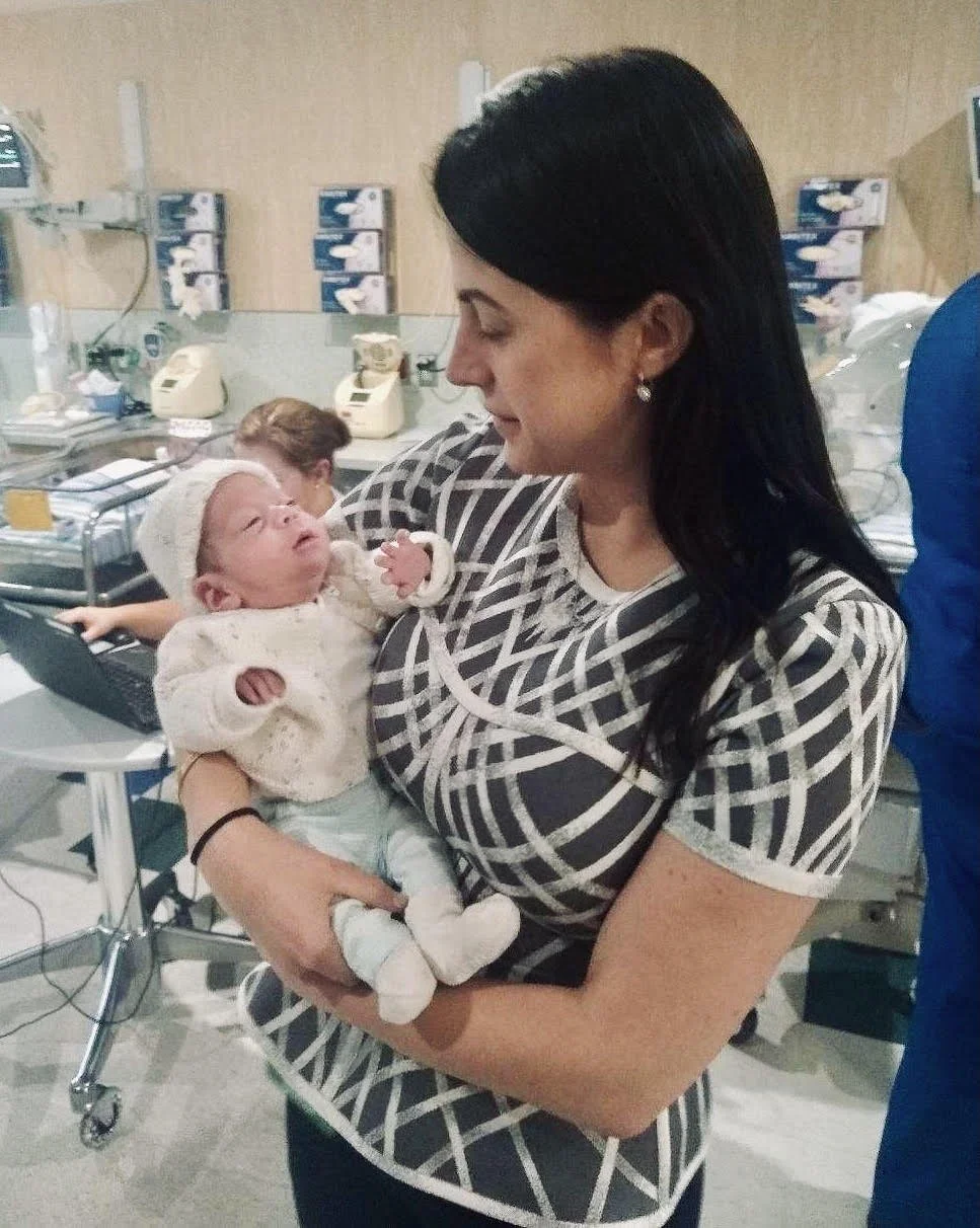 A woman holds a newborn baby in a hospital setting. The woman is looking at the baby, who is dressed in white. Medical equipment and staff are visible in the background.