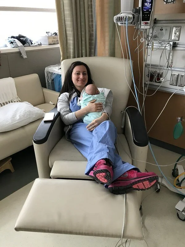 Hospital room with a woman in scrubs sitting in a chair, holding a newborn baby wrapped in a blanket; medical monitors and equipment are visible.