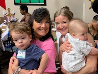 Two women holding young children in a room with other people in the background.