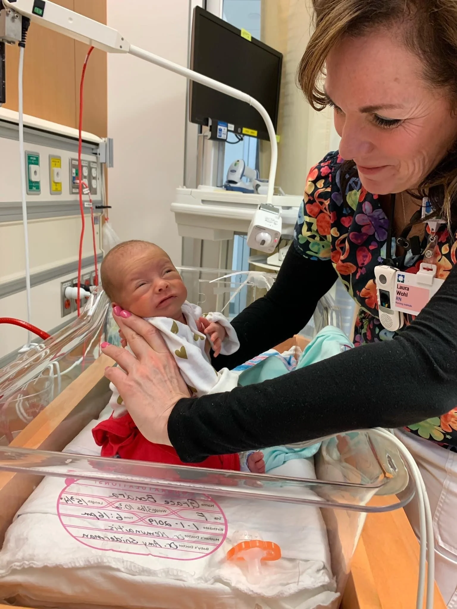 A woman holding a newborn baby in a hospital bassinet, smiling at the infant.