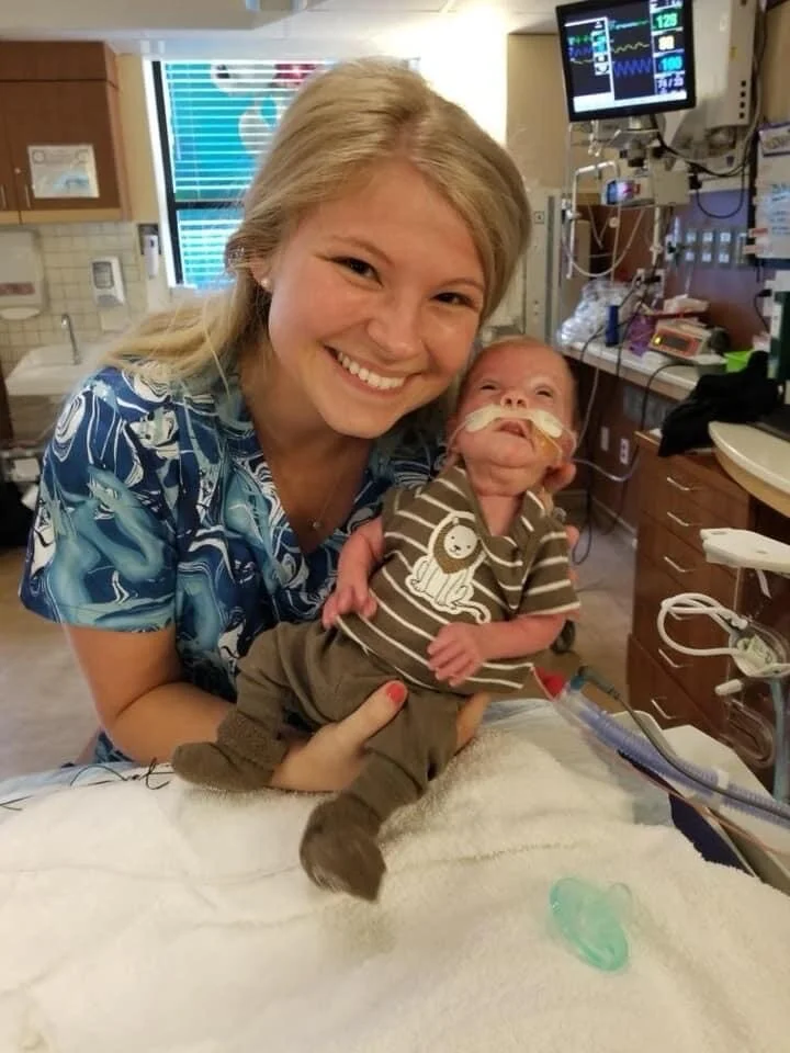 A smiling woman in hospital scrubs holding a baby with a feeding tube in a hospital room.