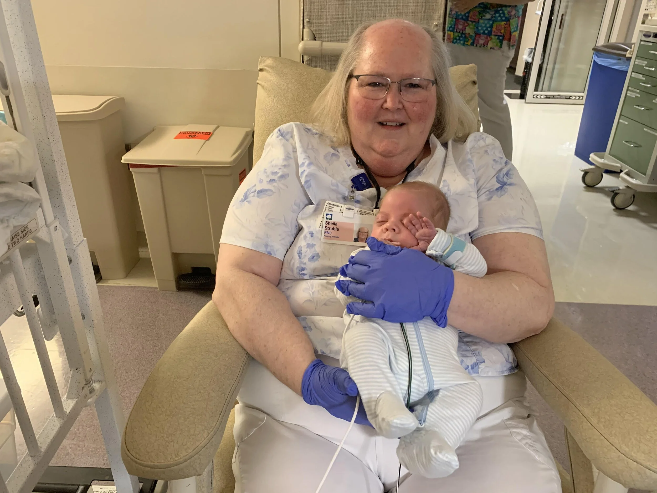 A woman in a hospital gown holding a newborn baby in her arms, both sitting on a beige hospital chair. The woman is smiling, wearing glasses and blue gloves. The baby is dressed in a striped onesie and is sleeping.
