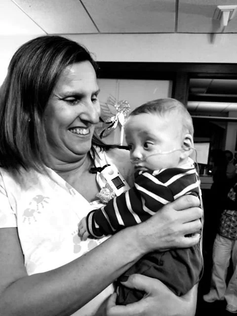 A woman holding a young boy with a nasal oxygen tube, both smiling at each other in an indoor setting.